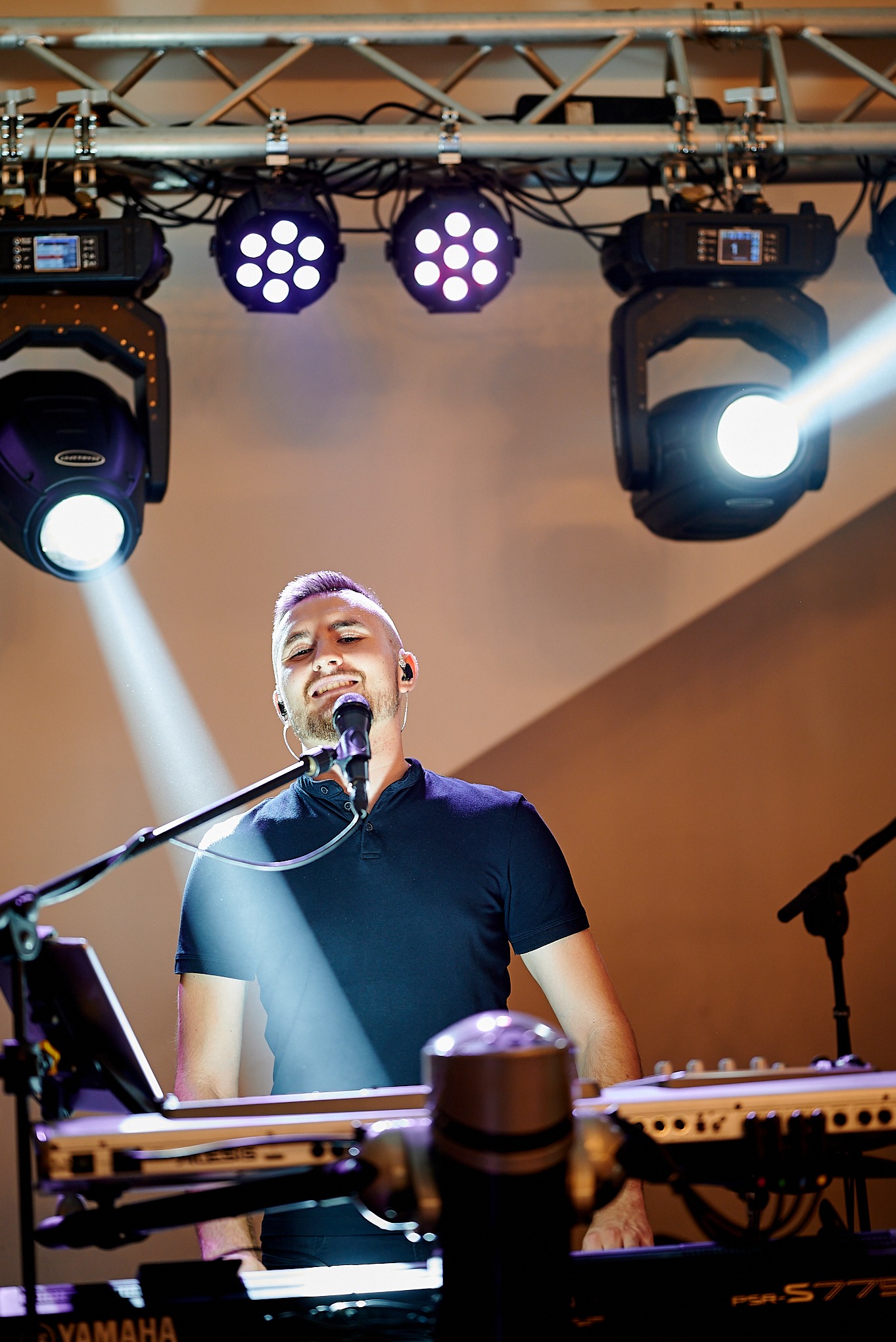 Wokalista z mikrofonem i słuchawkami na uszach, grający na keyboardzie Yamaha PSR-S775, oświetlony reflektorami punktowymi