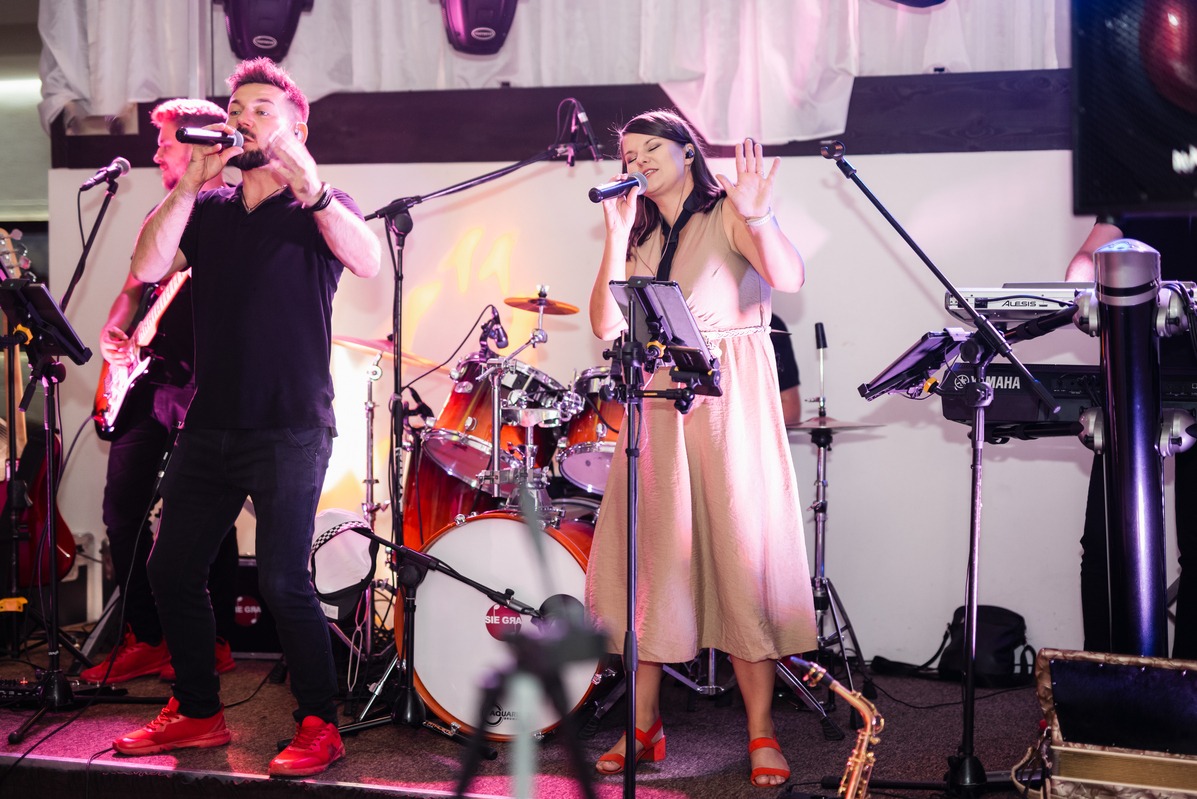 Zespół muzyczny podczas występu na scenie: wokalista w czarnej koszulce i czerwonych butach, wokalistka w beżowej sukience, perkusja, gitara, saksofon i klawisze.