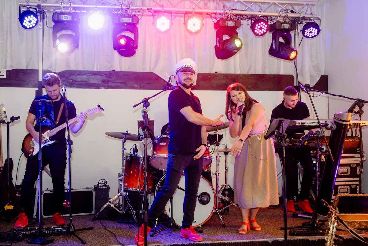 Zespół muzyczny na scenie podczas występu: gitarzysta gra na gitarze, wokalistka śpiewa do mikrofonu, klawiszowiec gra na keyboardzie, a perkusista siedzi za perkusją, wszyscy ubrani w czarne...