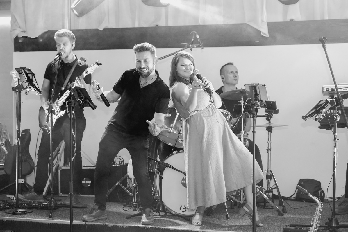 Czarno-białe zdjęcie zespołu muzycznego podczas występu na scenie: wokalistka w jasnej sukience z mikrofonem, gitarzysta z gitarą elektryczną i perkusista w tle.