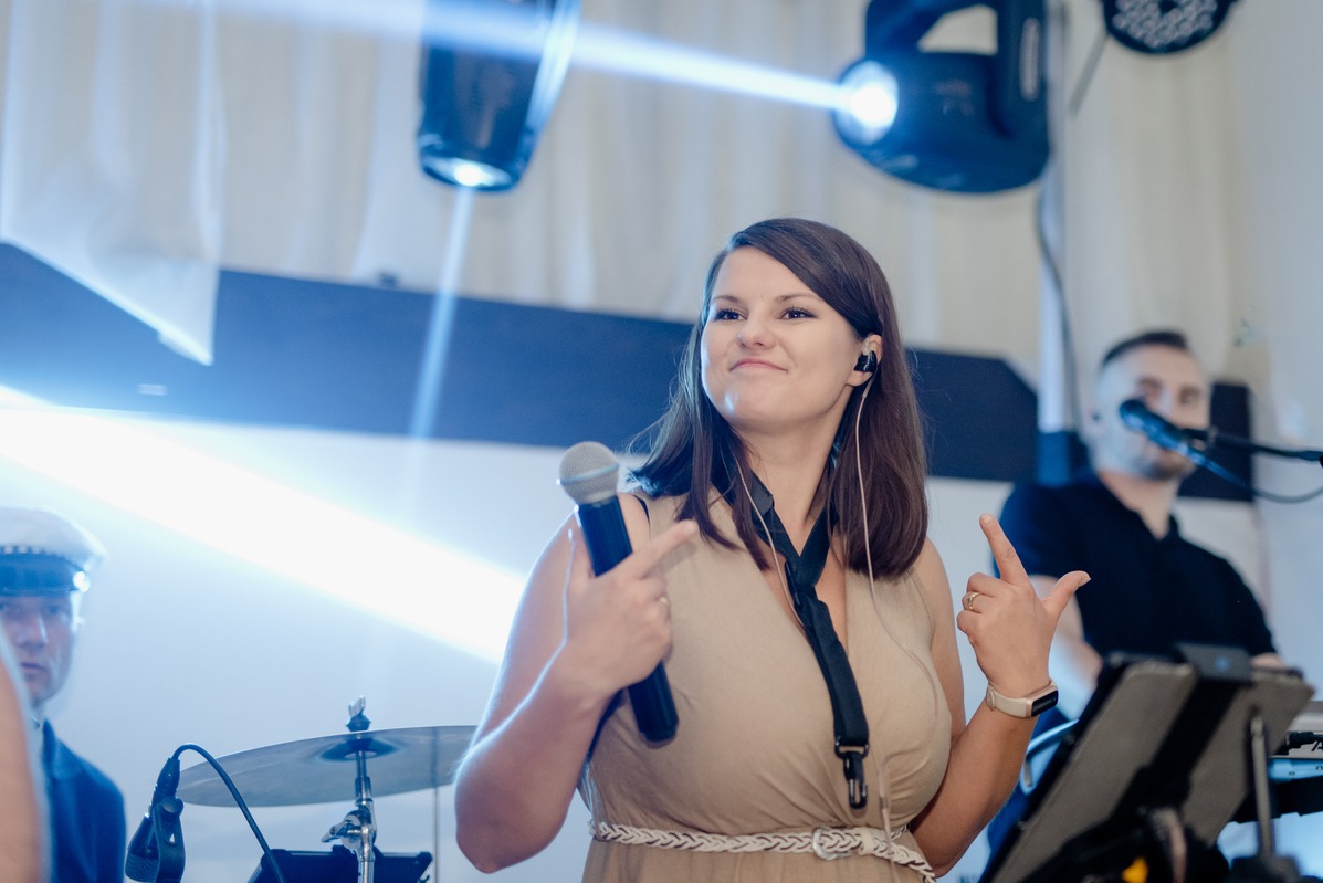 Uśmiechnięta wokalistka z mikrofonem w dłoni, w tle perkusja i klawiszowiec, oświetlenie sceniczne.