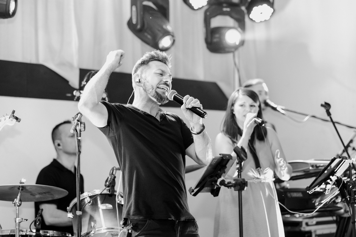 Czarno-białe zdjęcie zespołu muzycznego na scenie podczas występu, wokalista z podniesioną ręką śpiewa do mikrofonu, w tle perkusja i oświetlenie sceniczne.