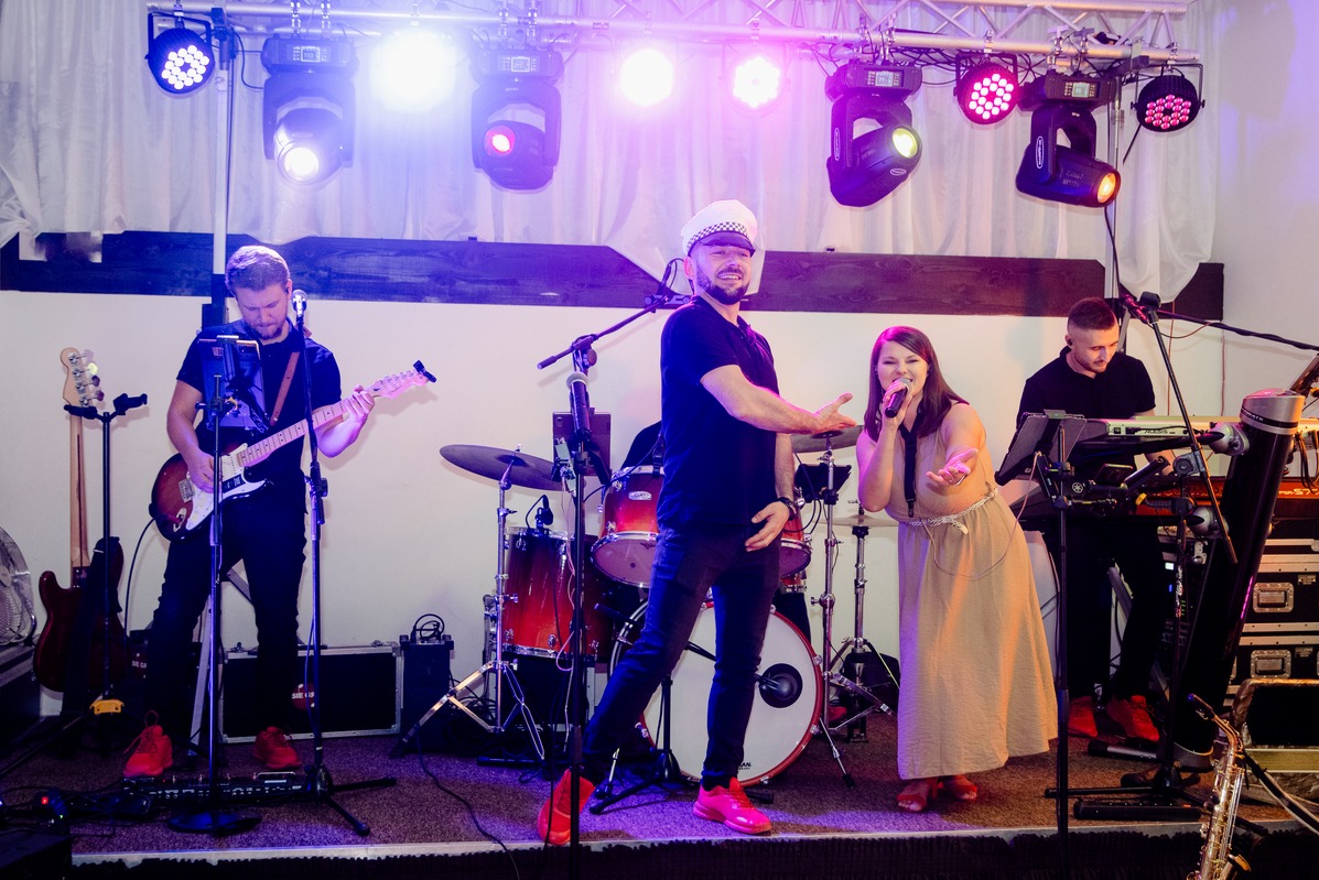 Zespół muzyczny na scenie podczas występu: gitarzysta, perkusista, wokalistka i klawiszowiec w czerwonych butach pod reflektorami.