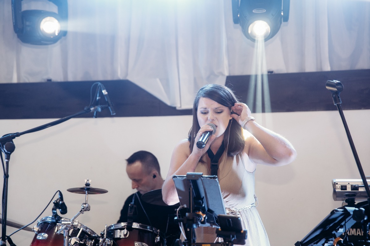 Wokalistka śpiewa do mikrofonu na scenie podczas występu, w tle perkusista i oświetlenie sceniczne.