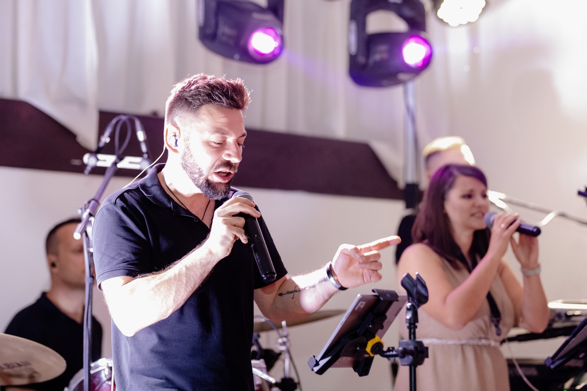 Zespół muzyczny w trakcie występu na scenie: wokalista z mikrofonem i tatuażem na przedramieniu, wskazujący palcem, obok wokalistka i perkusista, oświetlenie sceniczne.