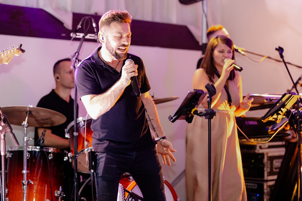 Wokalista z mikrofonem i wytatuowanym przedramieniem śpiewa na scenie z zespołem; perkusja i klawisze w tle.