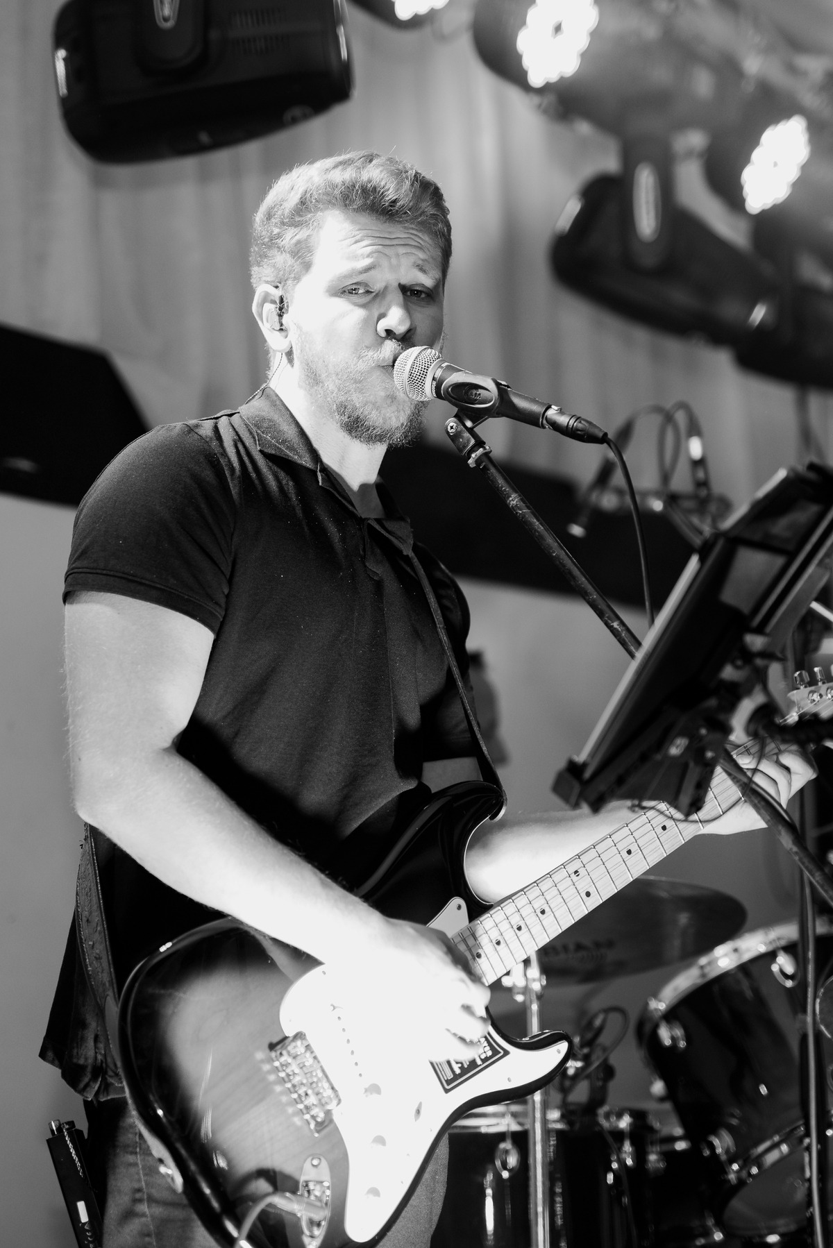 Czarno-białe zdjęcie mężczyzny z brodą śpiewającego do mikrofonu i grającego na elektrycznej gitarze podczas występu na scenie z oświetleniem i perkusją w tle.