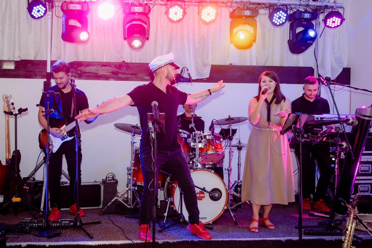 Zespół muzyczny w trakcie występu na scenie, wokalistka śpiewa do mikrofonu, lider zespołu w czapce marynarskiej gestykuluje, gitarzysta gra na gitarze, perkusista i klawiszowiec w tle, czerwone...