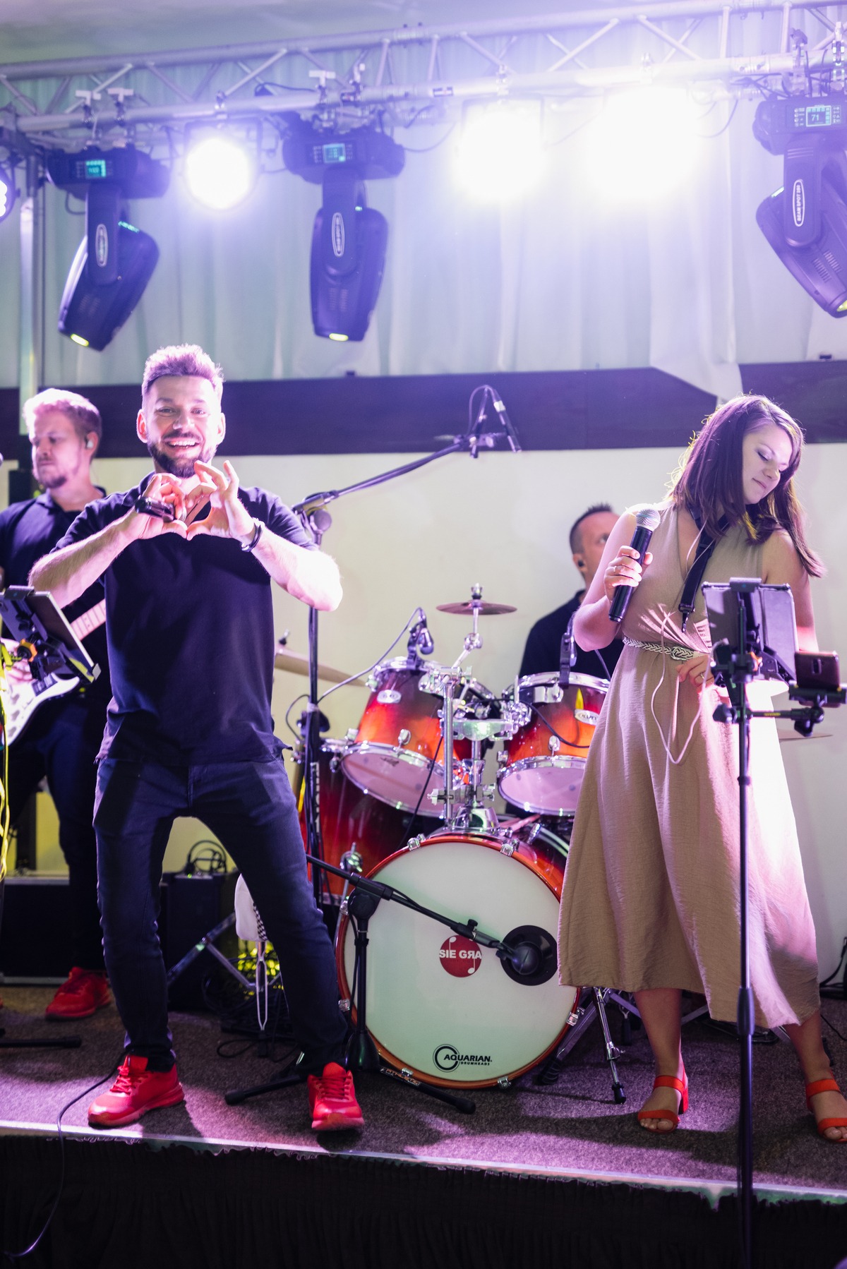 Występujący na scenie wokalista formuje dłońmi serce, obok wokalistka z mikrofonem, w tle perkusja i gitarzysta oświetleni reflektorami.