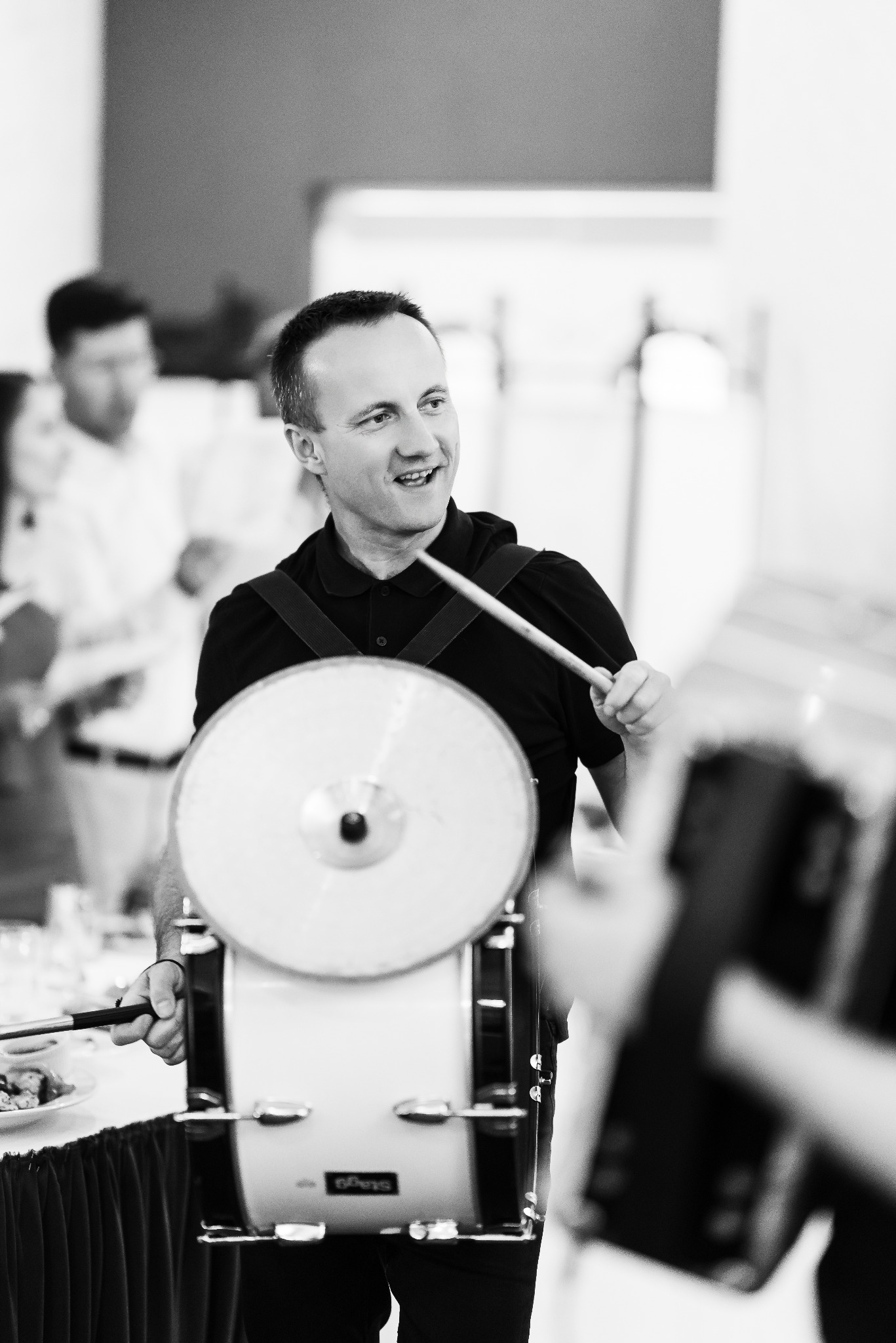 Czarno-białe zdjęcie uśmiechniętego perkusisty grającego na bębnie podczas występu, z widocznymi pałeczkami i fragmentami publiczności w tle.