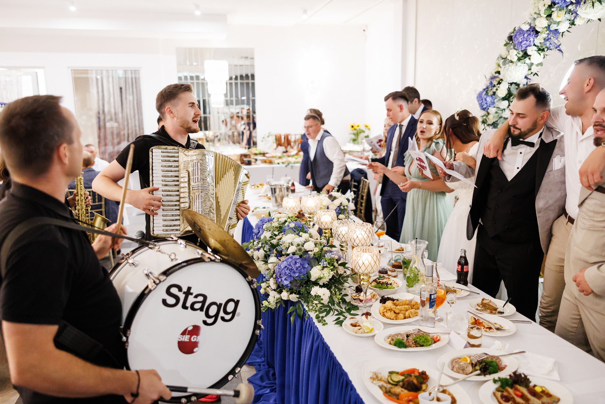 Zespół muzyczny grający na akordeonie i perkusji podczas wesela, goście śpiewają przy stole z dekoracjami kwiatowymi i jedzeniem. Jasne wnętrze sali weselnej.