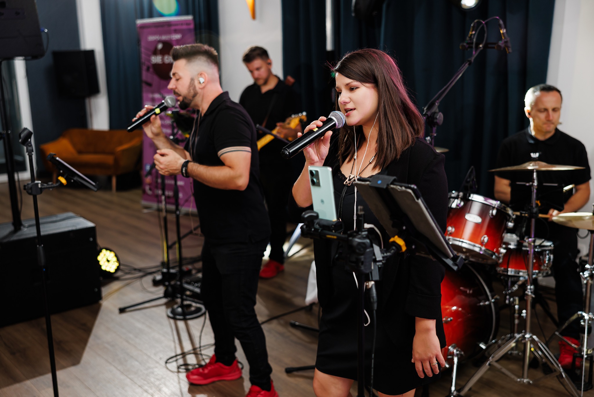 Zespół muzyczny w trakcie występu: wokalistka z mikrofonem, gitarzysta w tle, perkusista za zestawem. Oświetlenie sceniczne i profesjonalny sprzęt nagłośnieniowy.