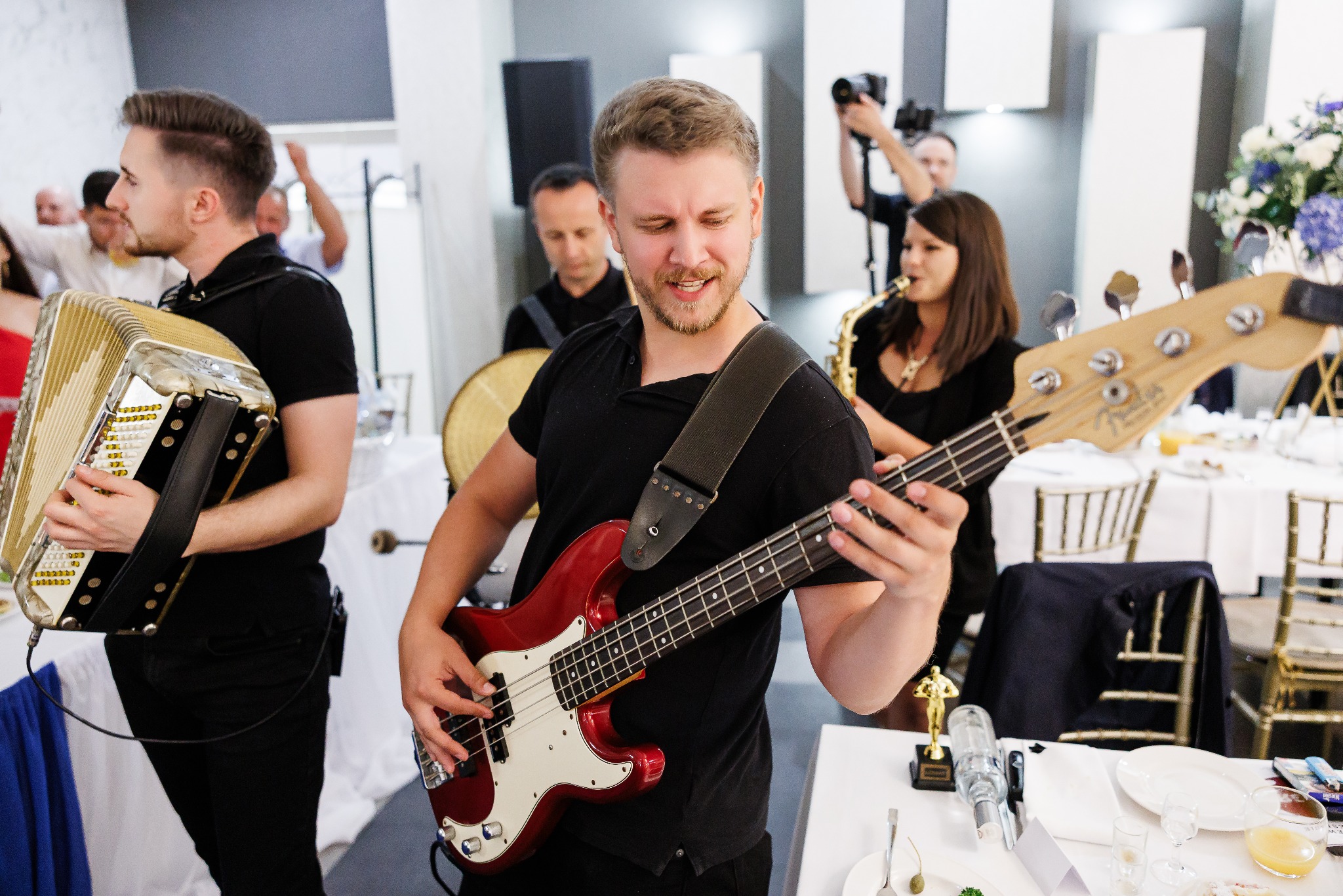Zespół muzyczny w akcji: basista gra na czerwonej gitarze basowej, obok akordeonista i saksofonistka. W tle perkusista i fotograf. Dekoracja stołu weselnego.
