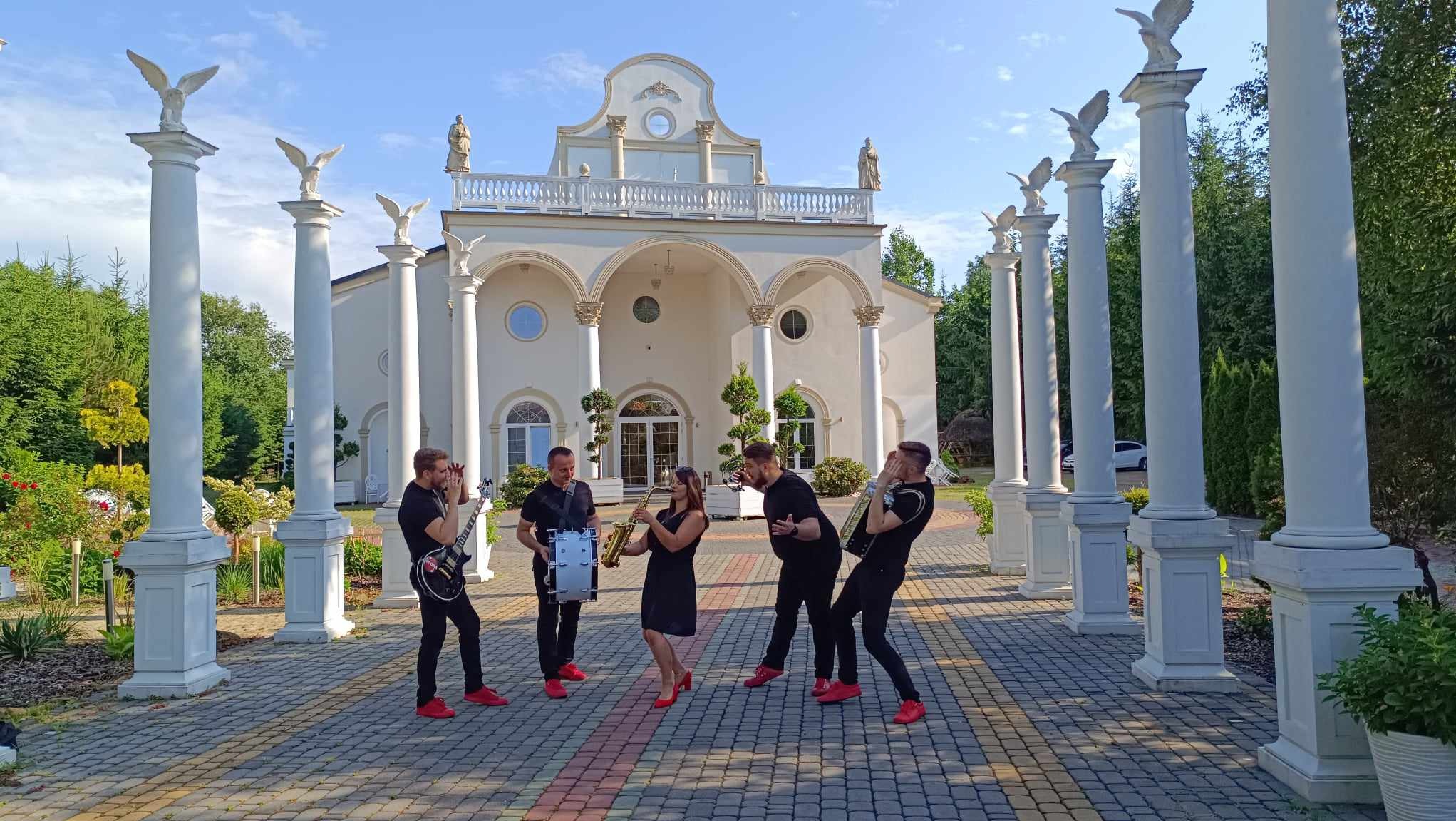 Zespół muzyczny w czarnych strojach i czerwonych butach gra na instrumentach przed białym budynkiem z kolumnami i rzeźbami orłów w słoneczny dzień.