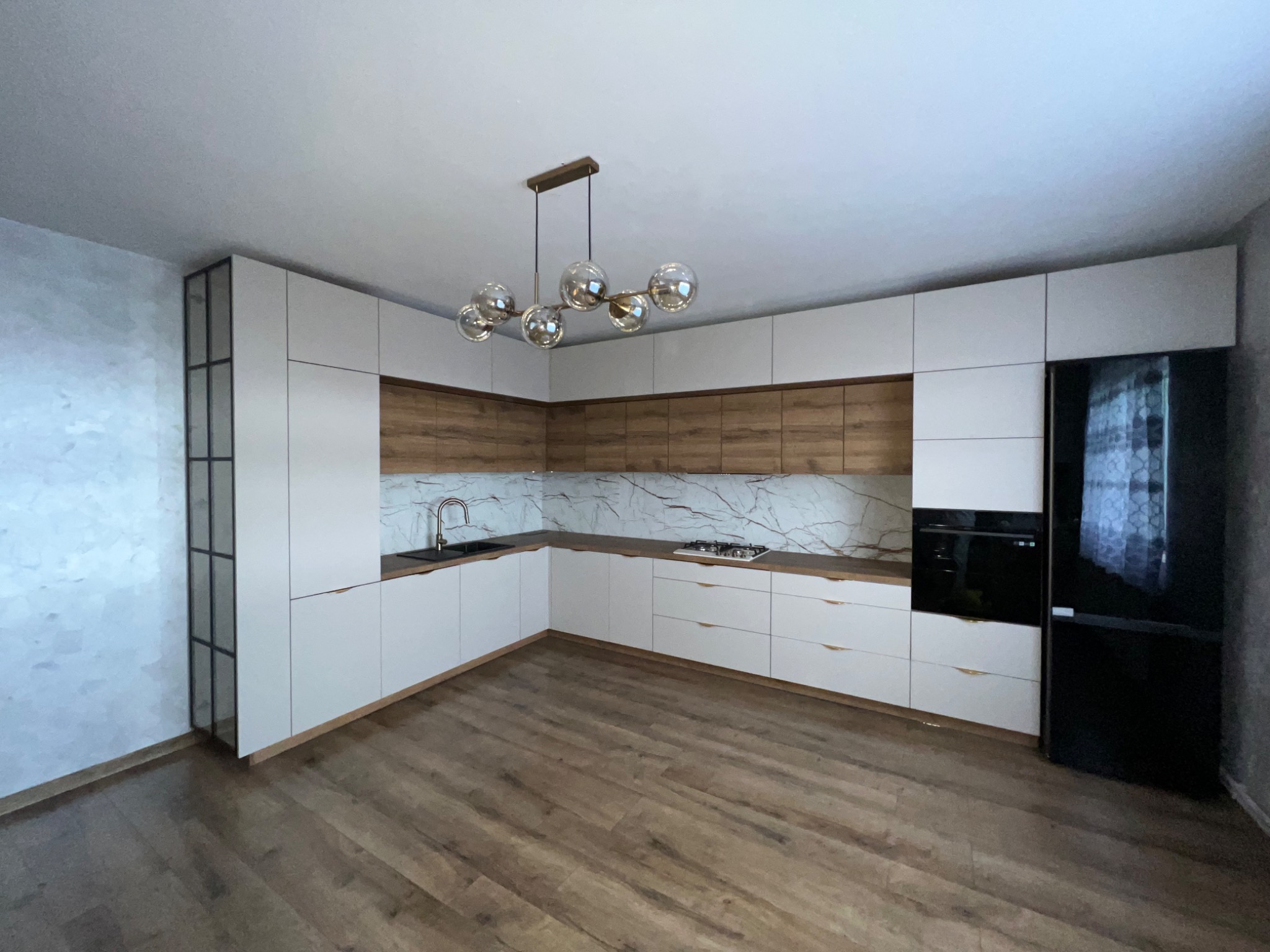 Nowoczesna kuchnia w odcieniach bieli i drewna, z marmurowym blatem, oświetlona dekoracyjną lampą wiszącą z kulistymi kloszami, widziana z perspektywy podłogi z paneli.