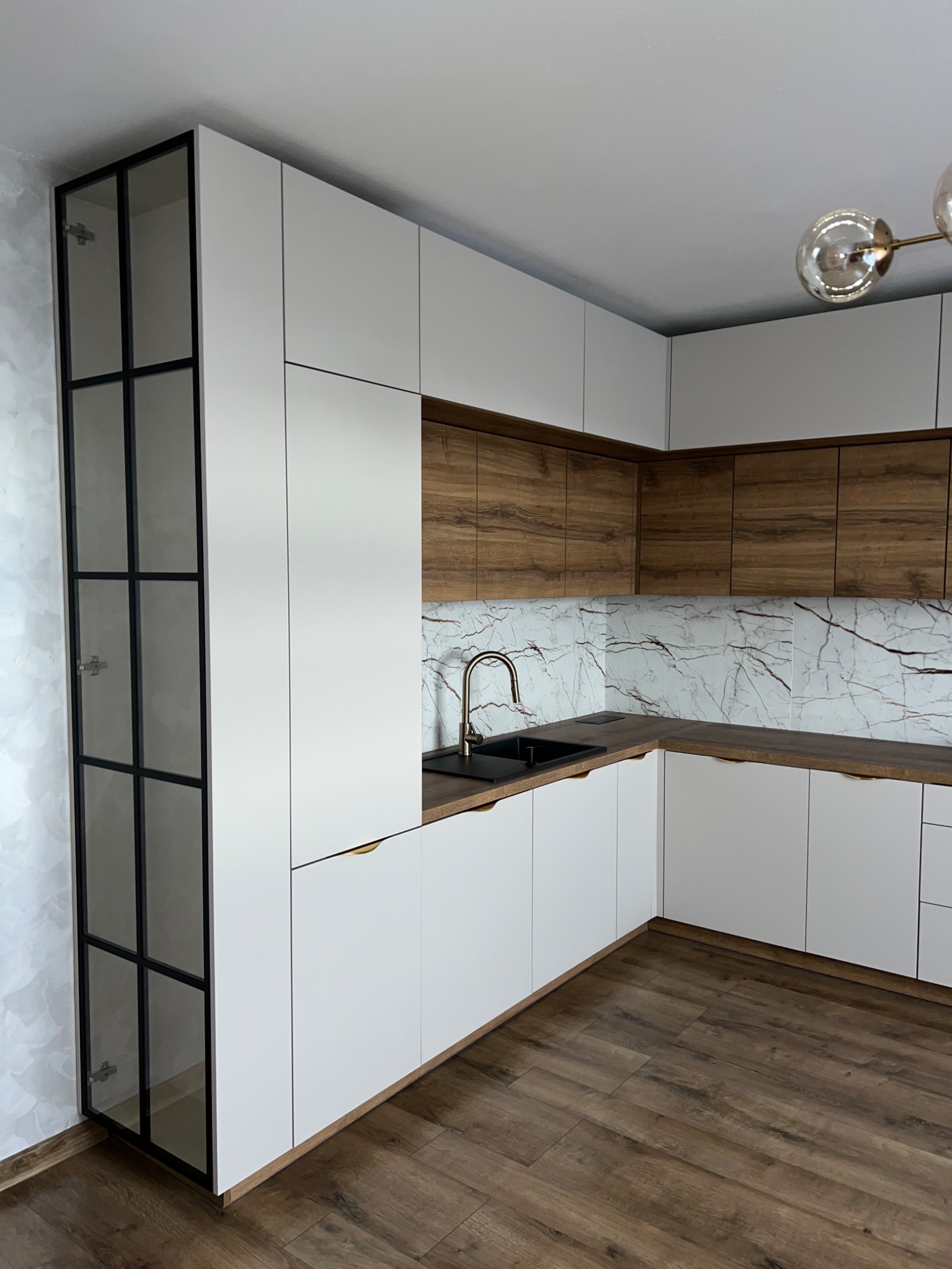 Nowoczesna kuchnia z białymi szafkami dolnymi i drewnopodobnymi szafkami górnymi, czarnym zlewem i złotą baterią, marmurowym backsplashiem oraz wysoką szafką z czarnymi szprosami i szklanymi...