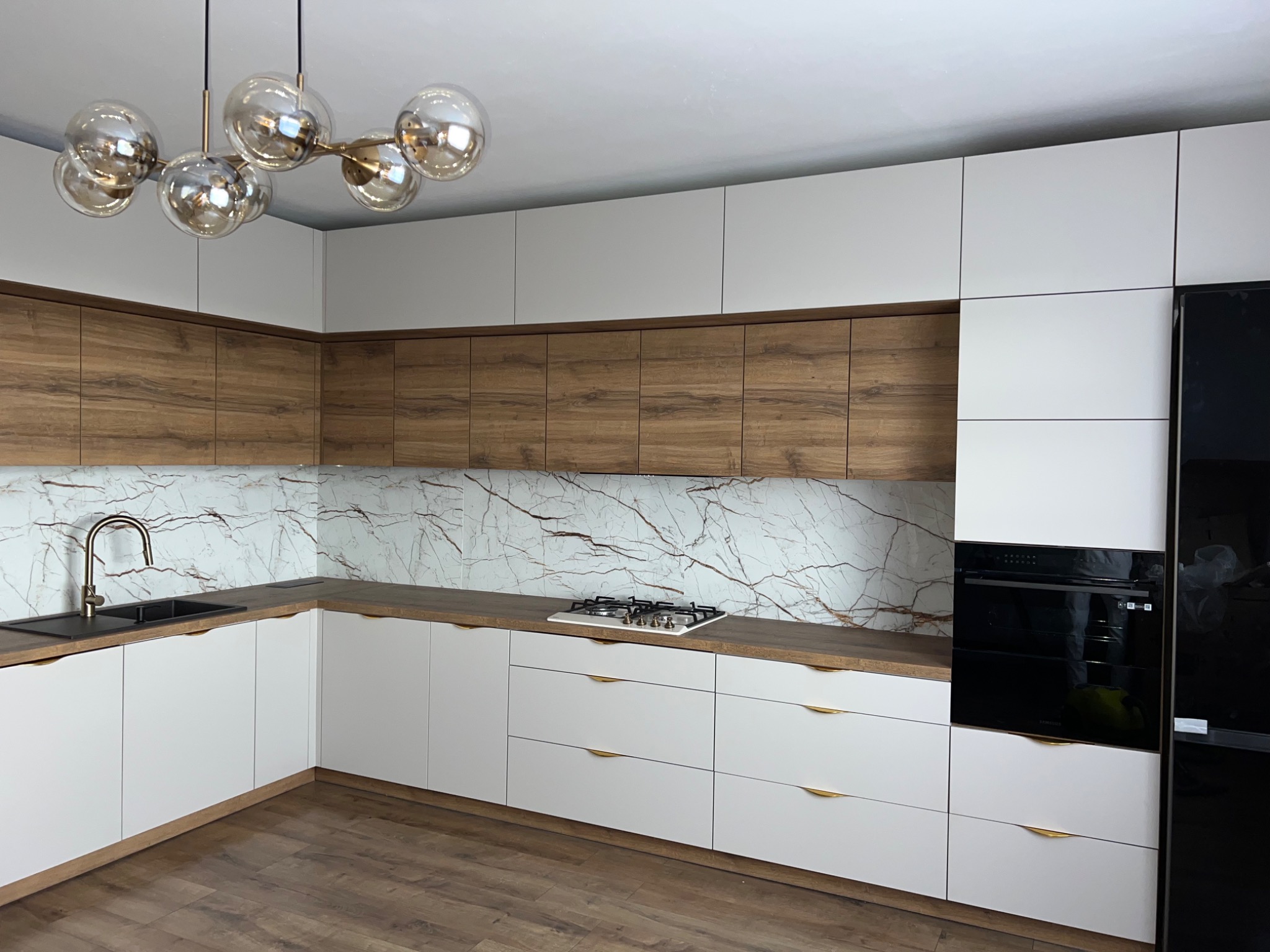 Nowoczesna kuchnia z białymi frontami szafek dolnych i górnych, drewnopodobnymi szafkami wiszącymi, blatem i marmurowym panelem nad blatem, oświetlona kulistą lampą.