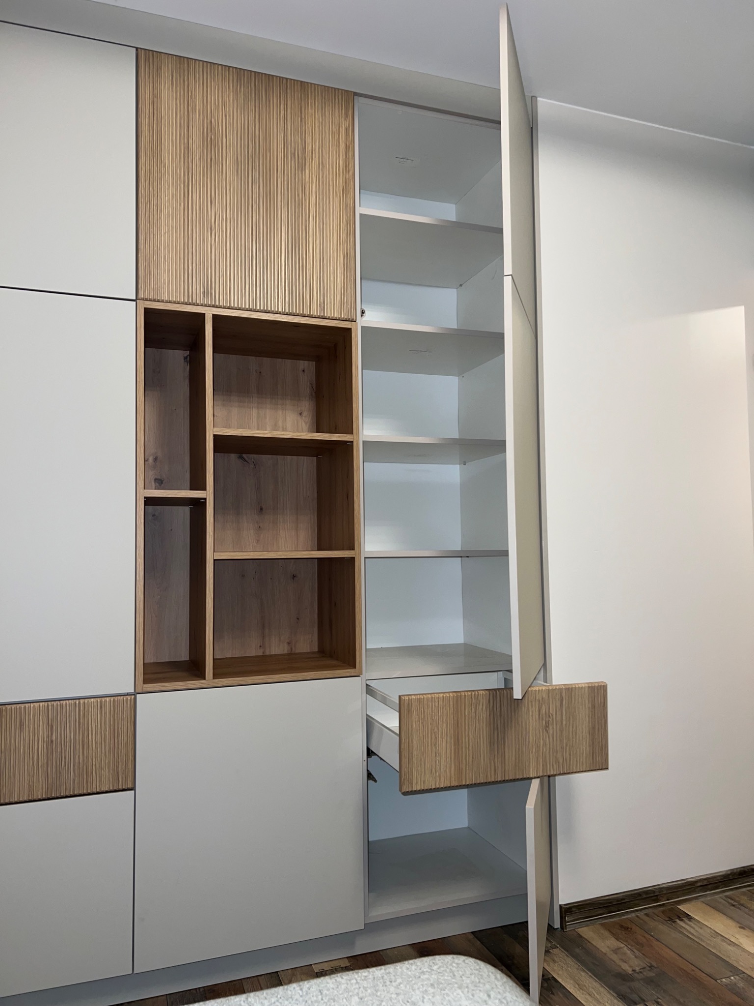 Nowoczesna szafa wnękowa z białymi frontami i elementami z drewna ryflowanego, otwarte półki i wysuwana szuflada z drewnianym frontem.