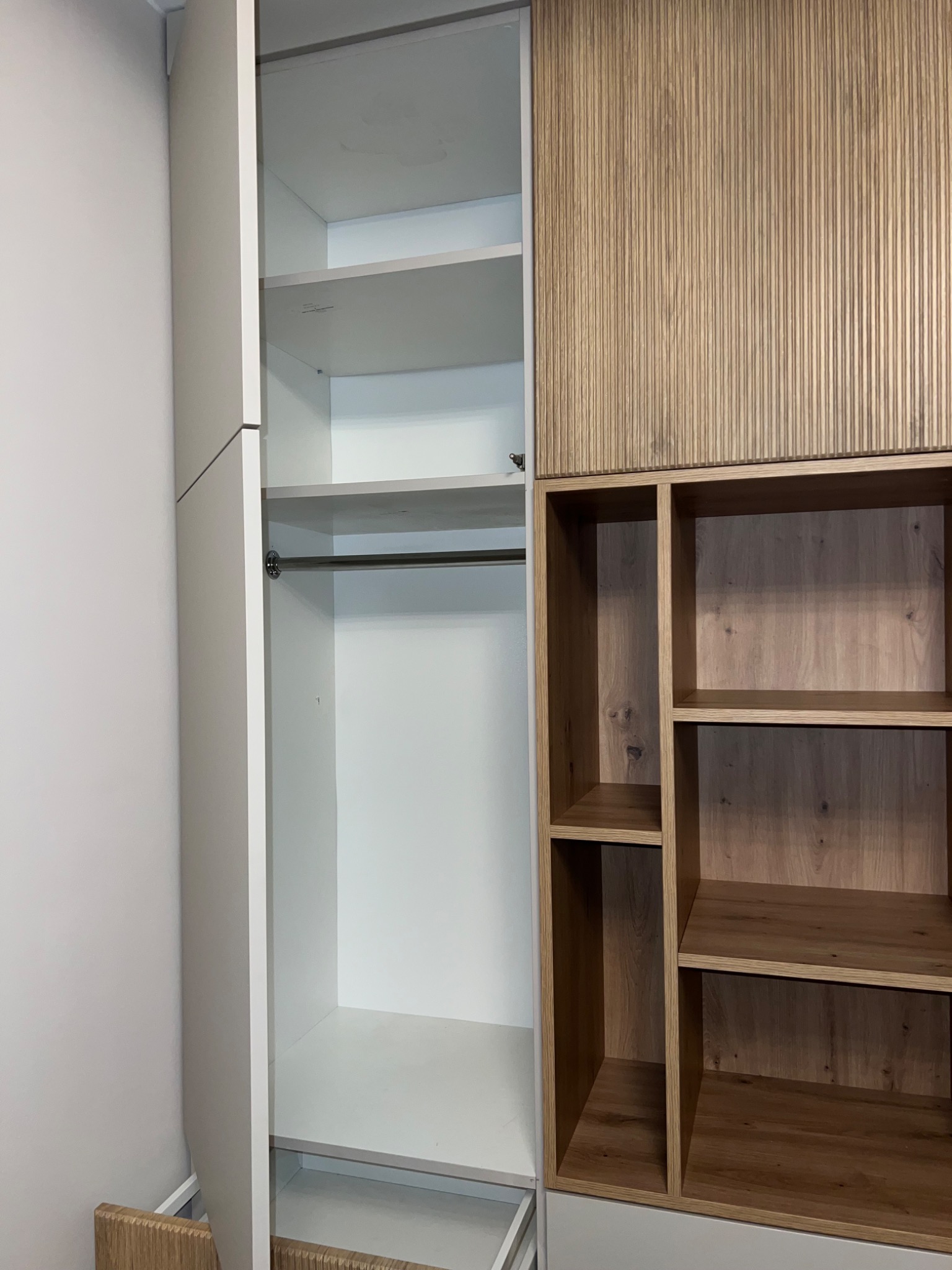 Wnętrze szafy z drzwiami w kolorze szarym, widoczne półki i drążek na ubrania, obok regał z półkami z drewna w odcieniach brązu, z dekoracyjnym frontem z pionowych listewek