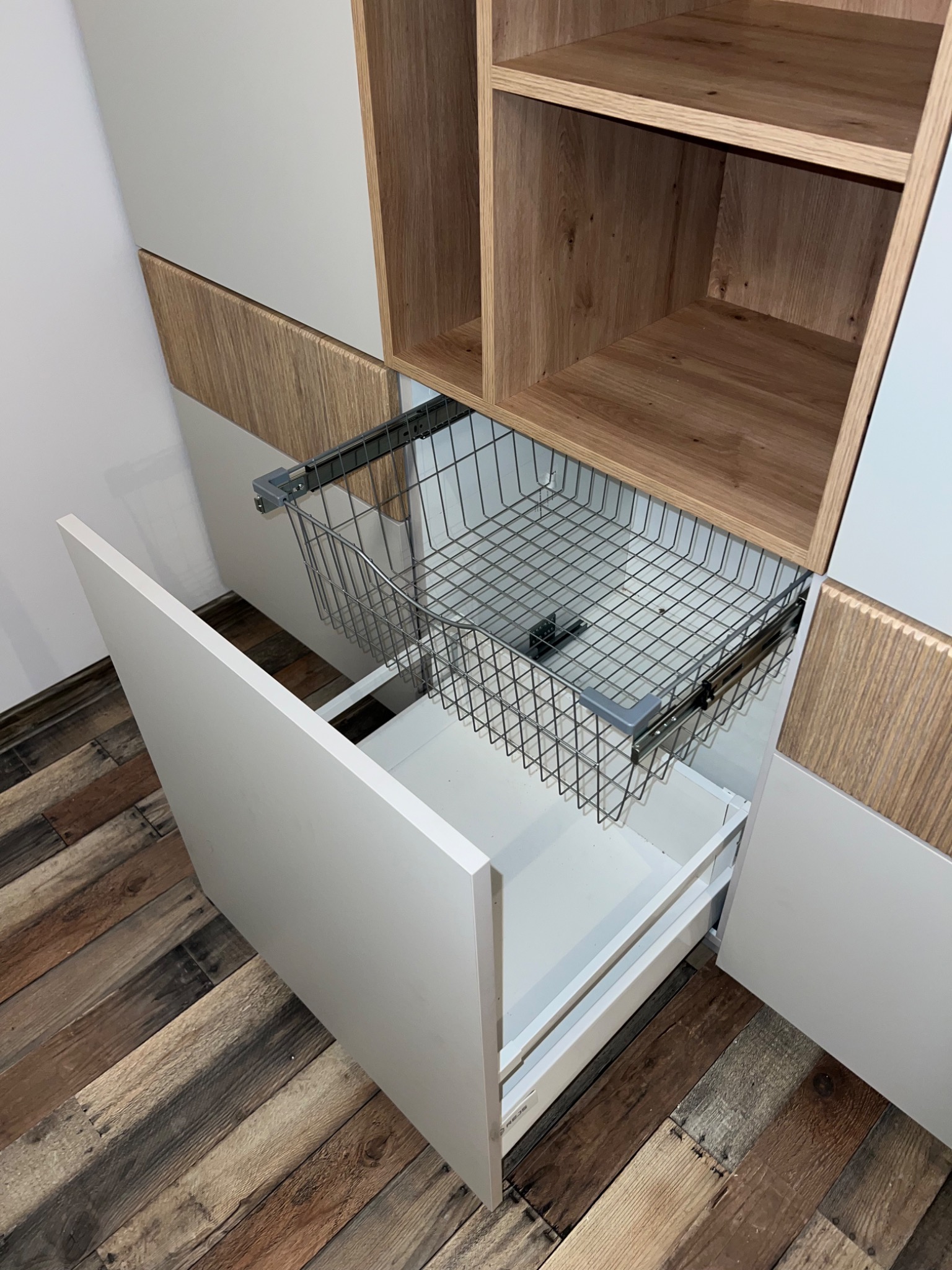 Wysuwana metalowa szuflada cargo w szafce z białymi frontami i drewnianymi akcentami, widok z góry na podłogę z desek w rustykalnym stylu.