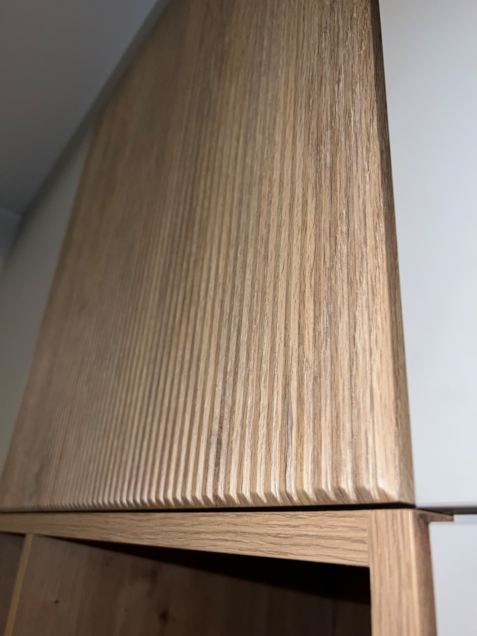 Fragment szafy z ryflowanym frontem z jasnego drewna, widoczne łączenie z białym panelem i półką.