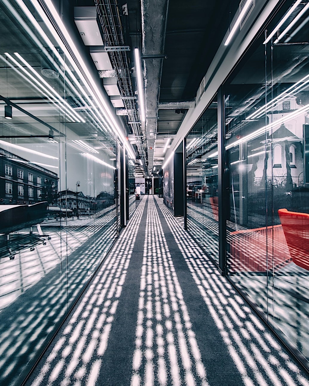 Długi korytarz biurowy z szklanymi ścianami, widoczne instalacje pod sufitem i odbicia budynków na szkle.