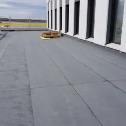 Płaski dach pokryty szarą papą termozgrzewalną na budynku z białymi ścianami i czarnymi oknami; w tle widoczne materiały budowlane i zabezpieczenia.