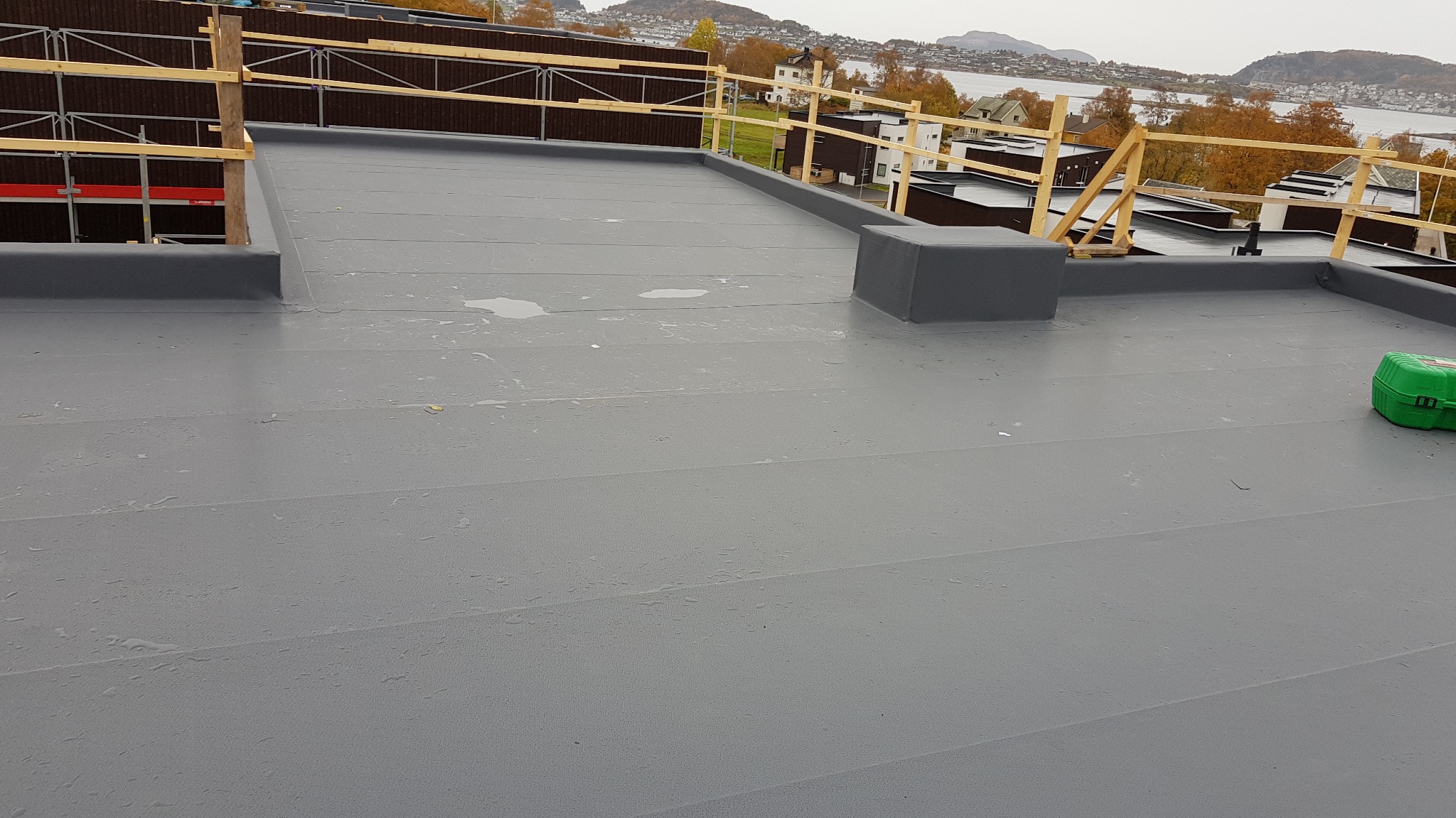 Szary, płaski dach pokryty papą z widocznymi śladami wilgoci, zabezpieczony drewnianą barierką, z zielonym pojemnikiem narzędziowym w prawym rogu i widokiem na zabudowania oraz zbiornik wodny w tle.