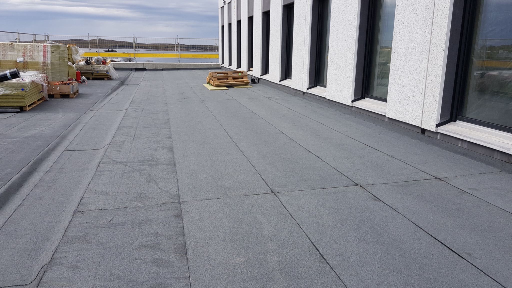Płaski dach pokryty szarą papą termozgrzewalną na budynku z białymi ścianami i czarnymi oknami; w tle widoczne materiały budowlane i zabezpieczenia.