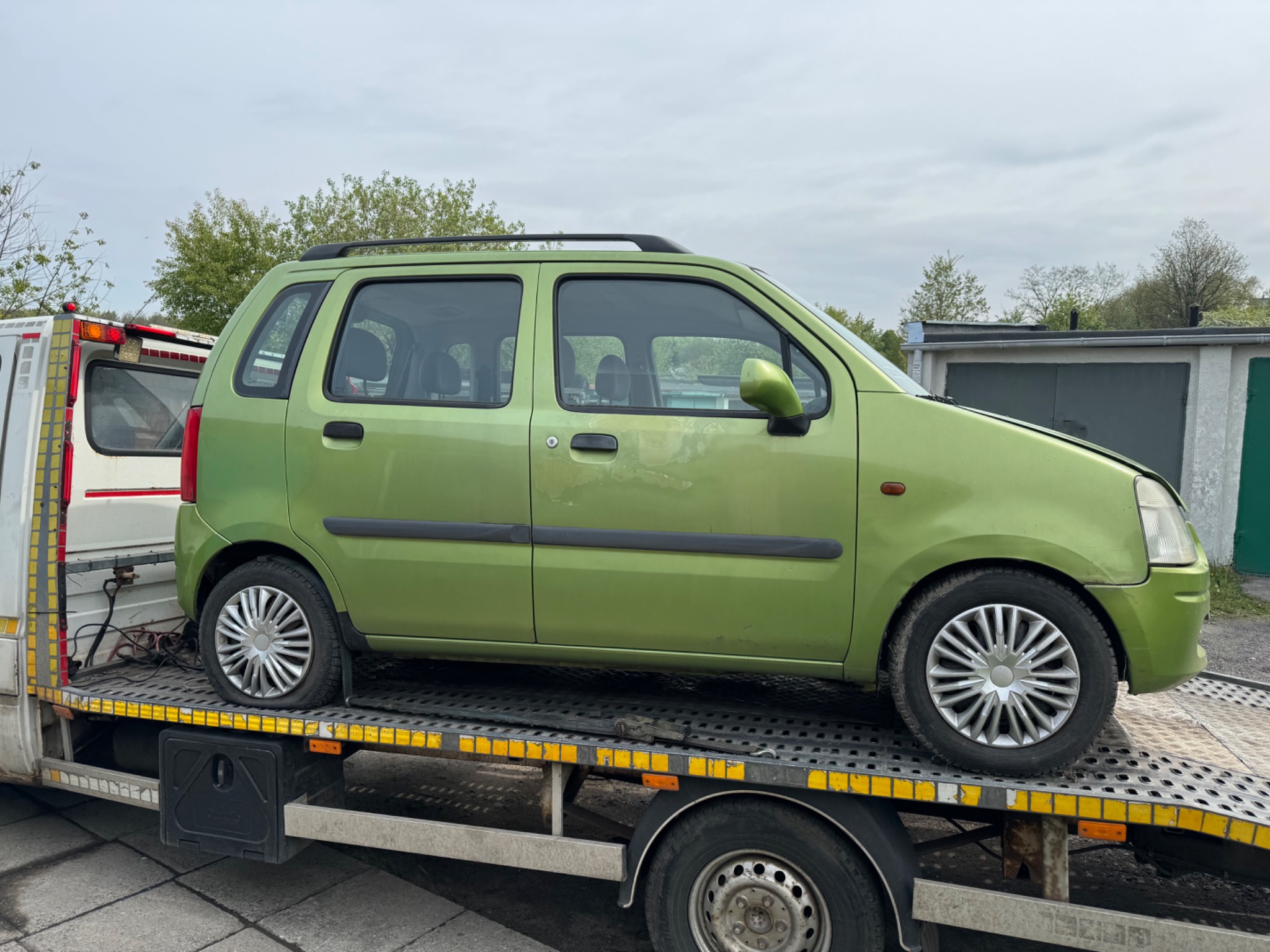Zielony samochód osobowy, z uszkodzonym przednim kołem, transportowany na lawecie auto pomocy, widok z boku.