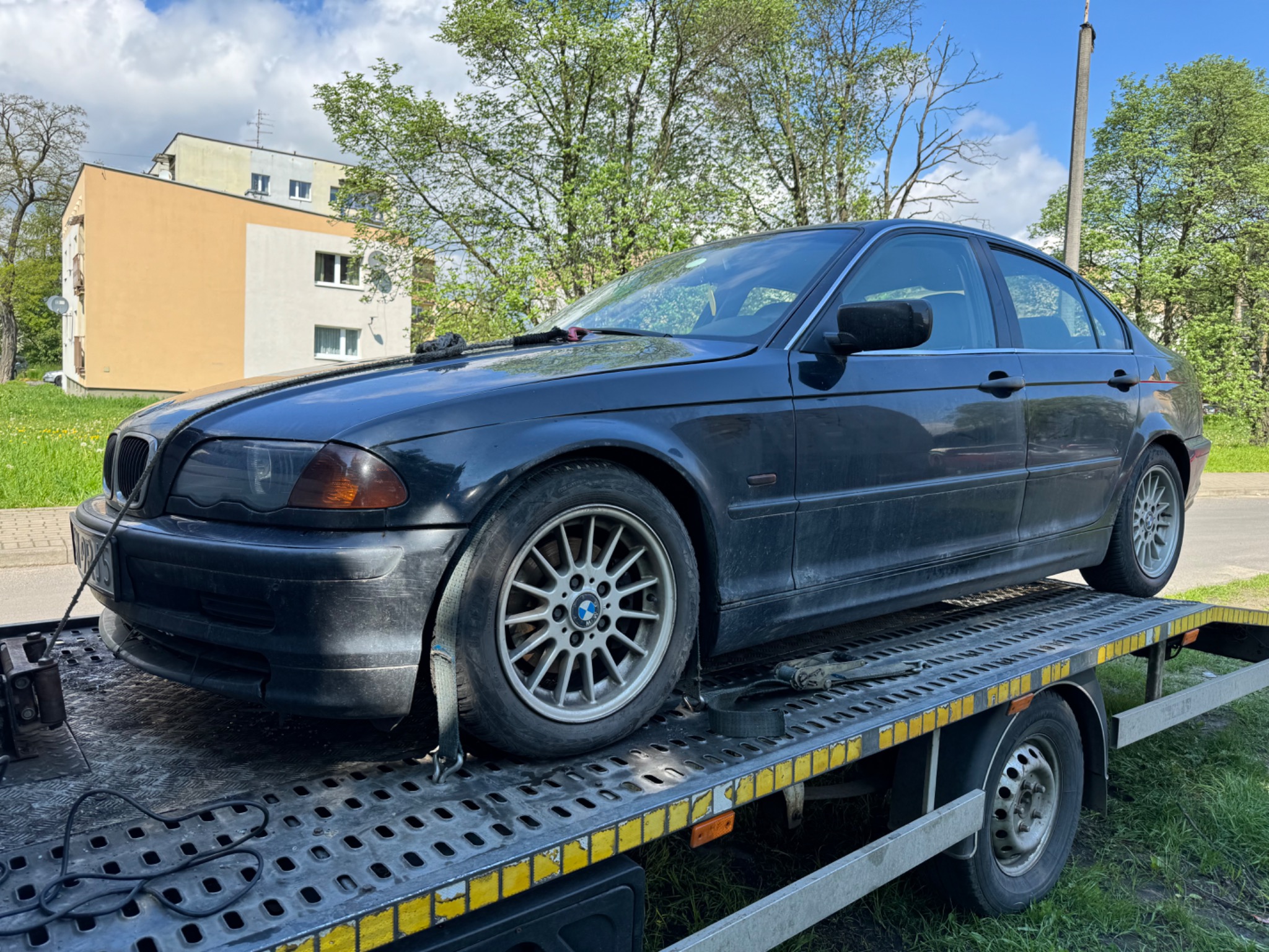 Czarny sedan BMW serii 3 (E46) zabezpieczony pasami transportowymi na platformie lawety holowniczej z widocznym budynkiem mieszkalnym w tle.
