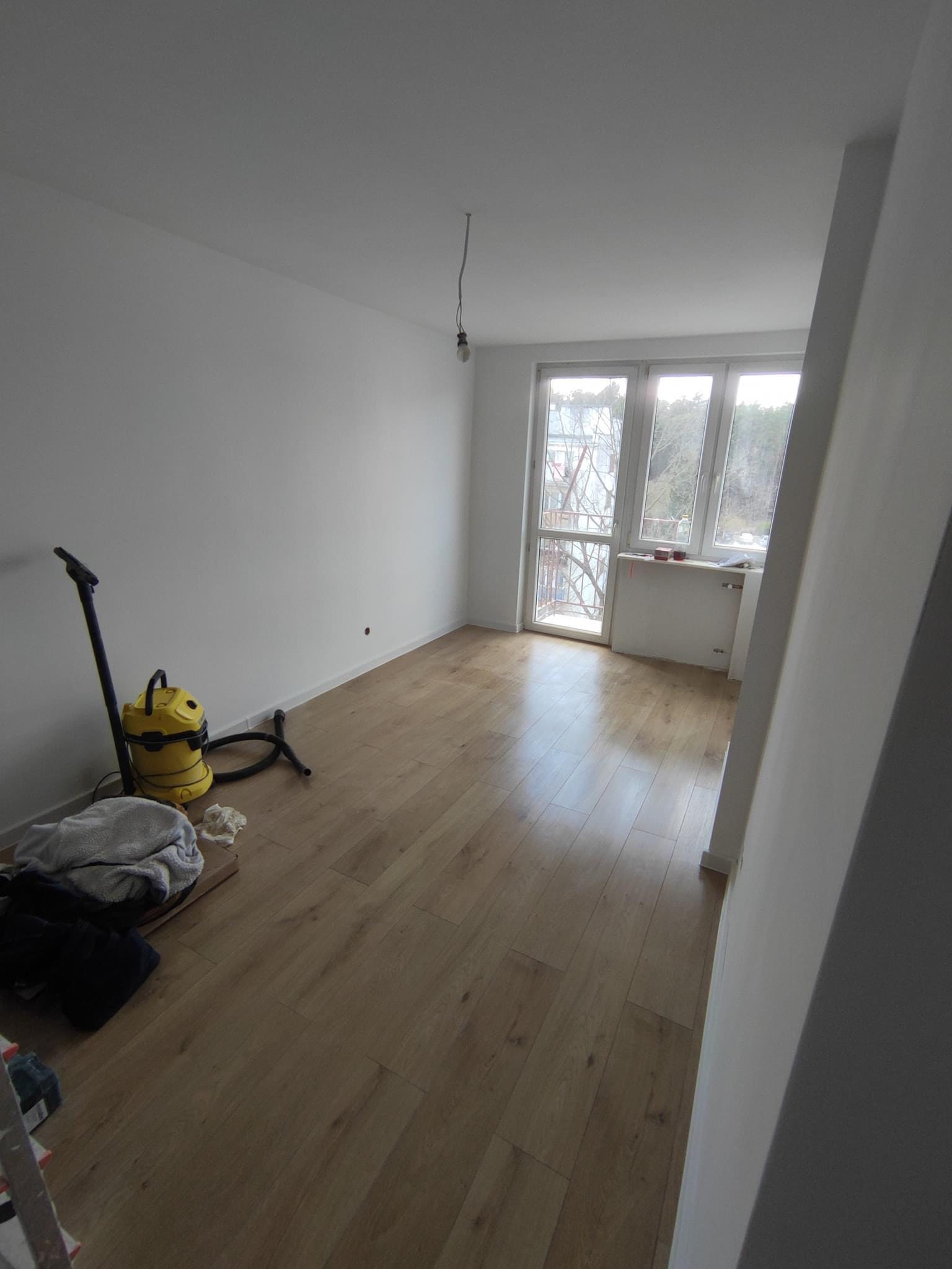 Pusty pokój po remoncie z jasnymi panelami podłogowymi, białymi ścianami i oknem balkonowym. Na podłodze stoi żółty odkurzacz przemysłowy oraz leżą szmaty i karton.
