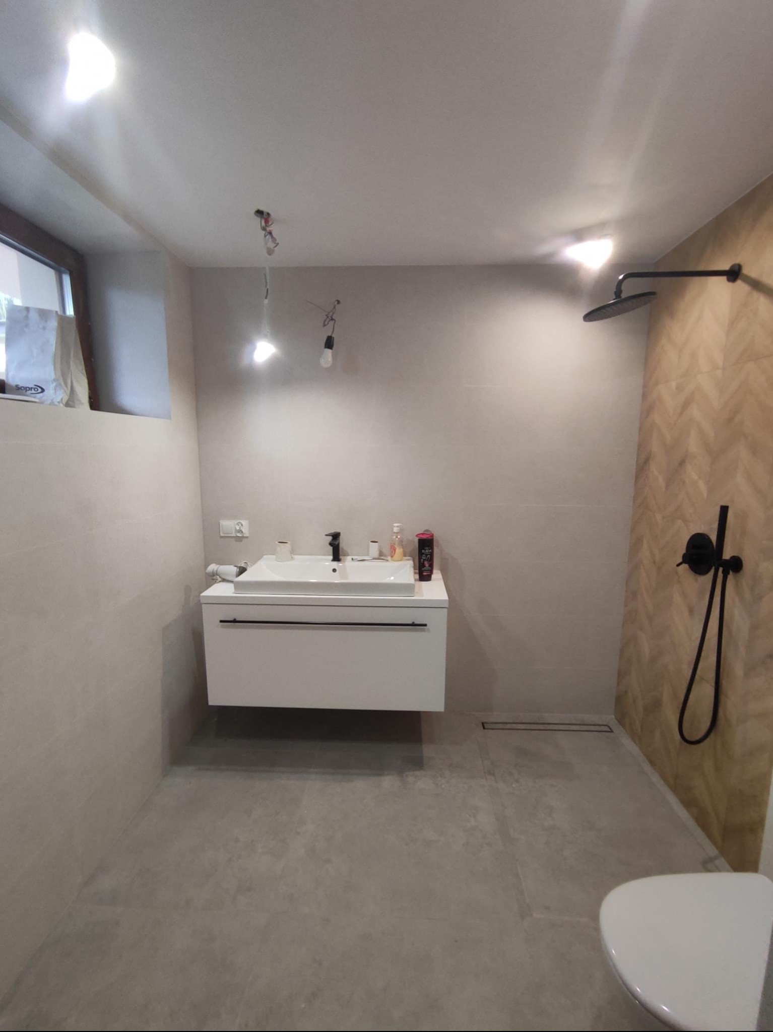 Nowoczesna łazienka z szarymi płytkami i białymi meblami, czarna armatura, widoczne niedokończone podłączenie oświetlenia sufitowego.