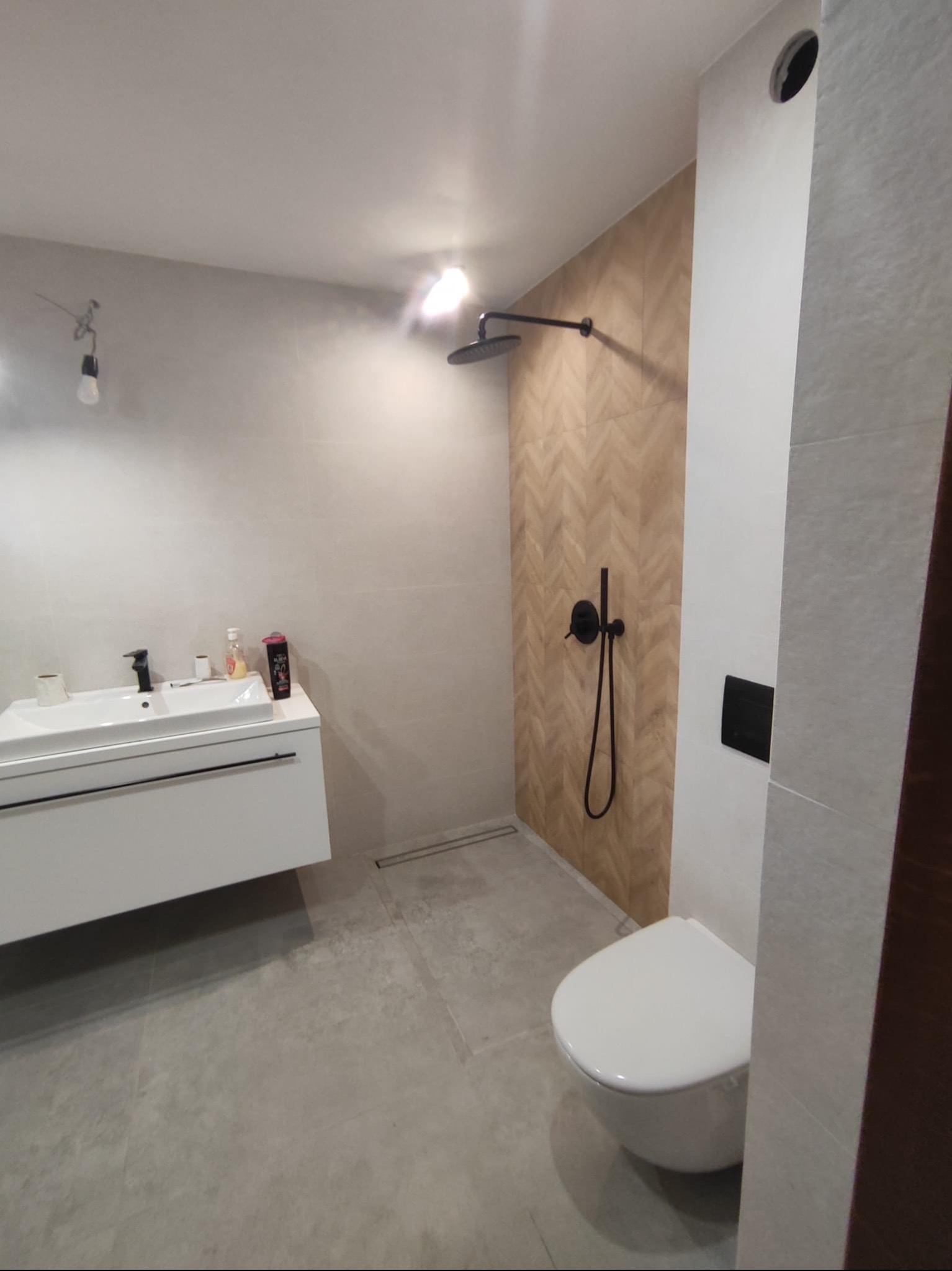 Nowoczesna łazienka z białą umywalką nablatową, czarną armaturą, deszczownicą i toaletą typu Geberit, wykończona płytkami w odcieniach szarości i beżu ułożonymi we wzór jodełki.