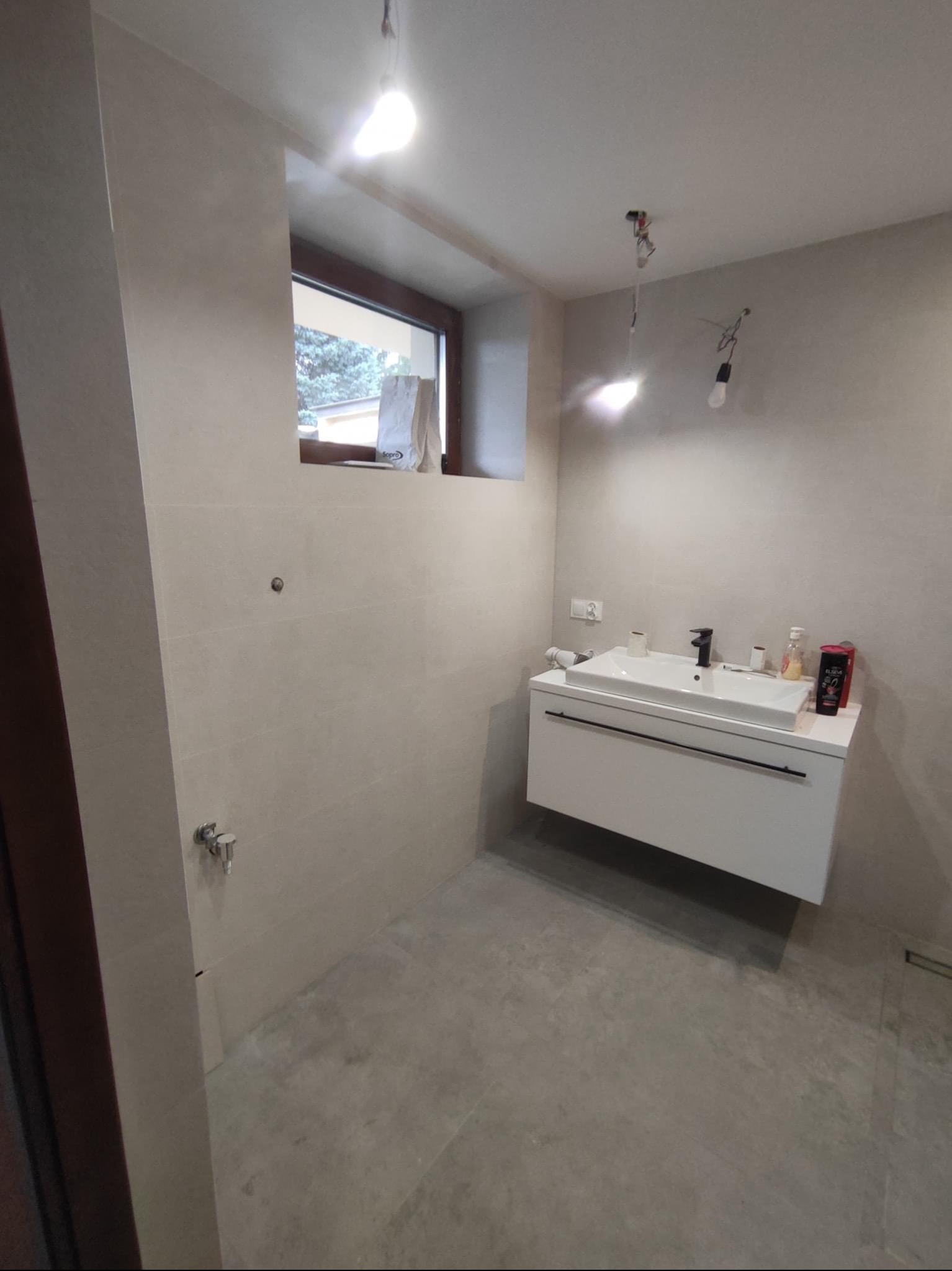 Wykończona łazienka w odcieniach beżu z wiszącą szafką z umywalką, szarą podłogą i oknem z widokiem na zewnątrz; widoczne niedokończone podłączenie oświetlenia sufitowego.