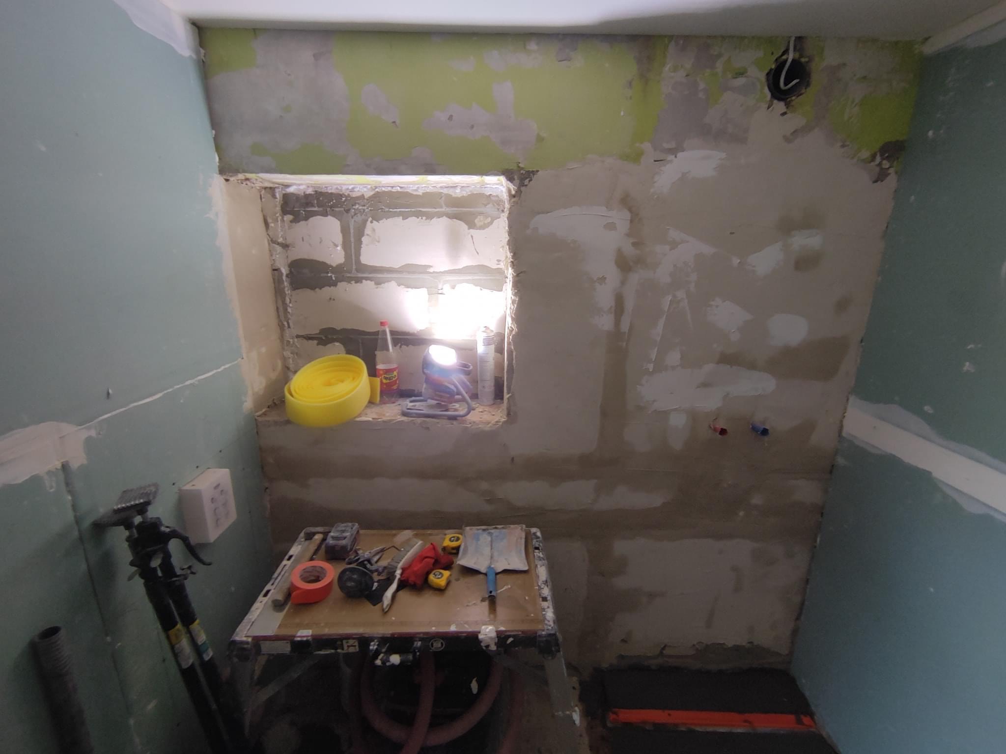Wnętrze łazienki w trakcie remontu, ściany częściowo pokryte gładzią, widoczne narzędzia na stole roboczym i fragment płyty karton-gips na ścianie, otwór w ścianie z cegieł.