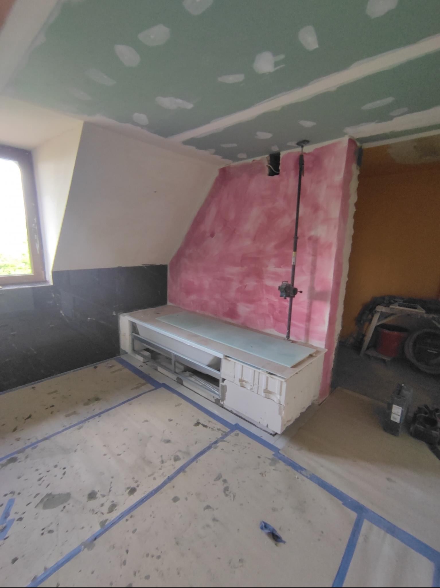 Wnętrze w trakcie prac remontowych: sufit z płyt kartonowo-gipsowych z widocznymi łączeniami, ściana z fragmentami różowej farby, czarne płytki przy oknie i przygotowany pod zabudowę niski element...