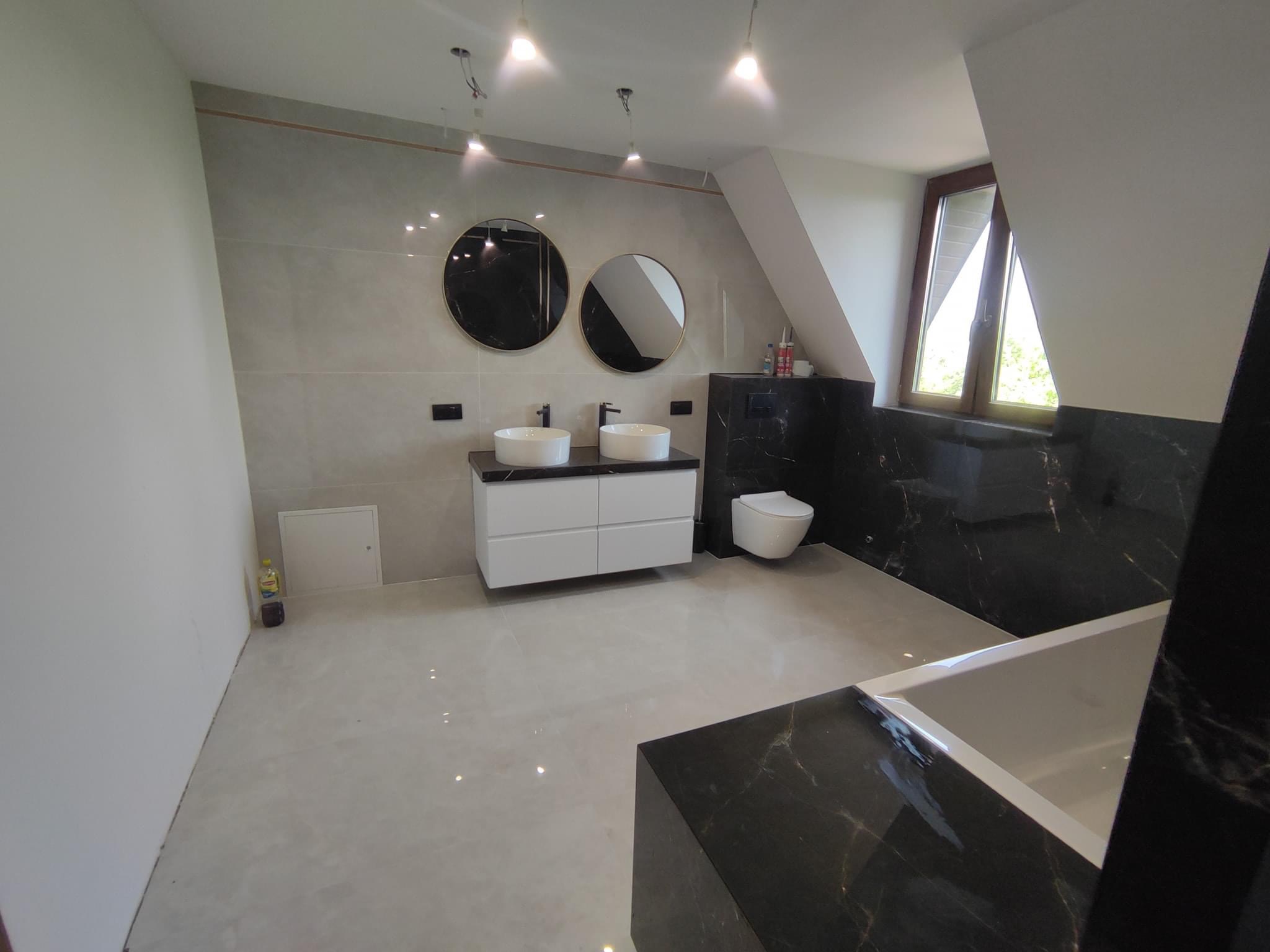 Nowoczesna łazienka na poddaszu z szarymi płytkami, czarnym marmurem, okrągłymi lustrami w złotych ramach, białą szafką z umywalkami nablatowymi i wiszącą toaletą.