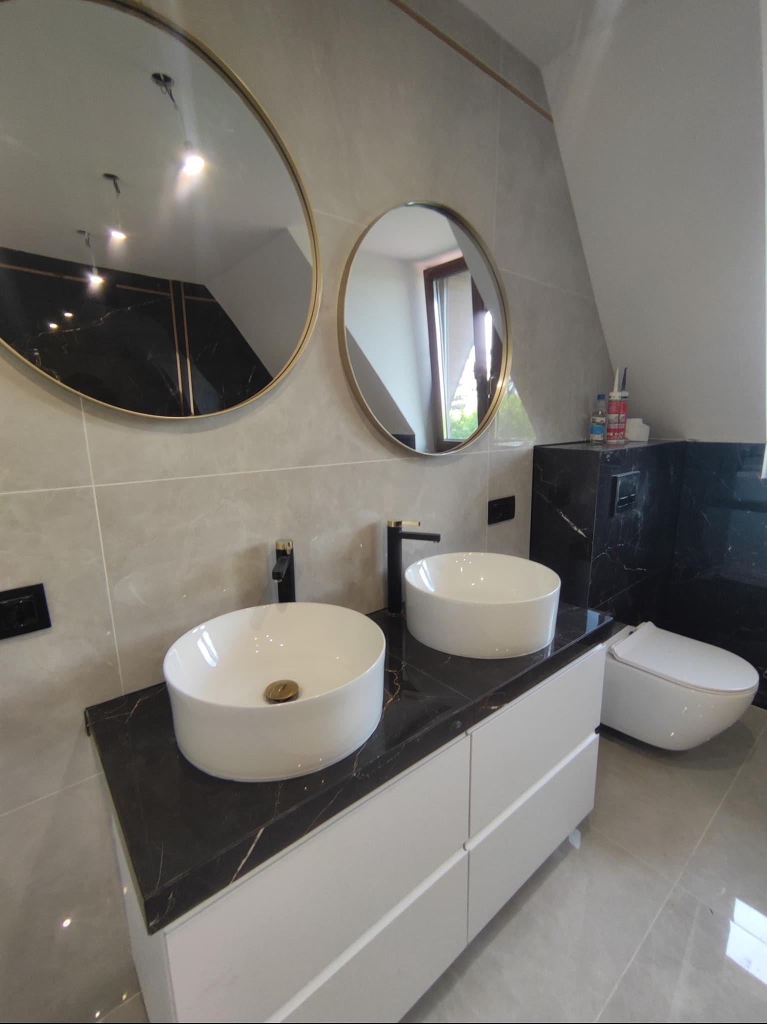 Nowoczesna łazienka na poddaszu z dwiema okrągłymi umywalkami nablatowymi, czarnym blatem z marmurowym wzorem, białymi szafkami pod umywalkami, dwoma okrągłymi lustrami w złotych ramach i szarą...
