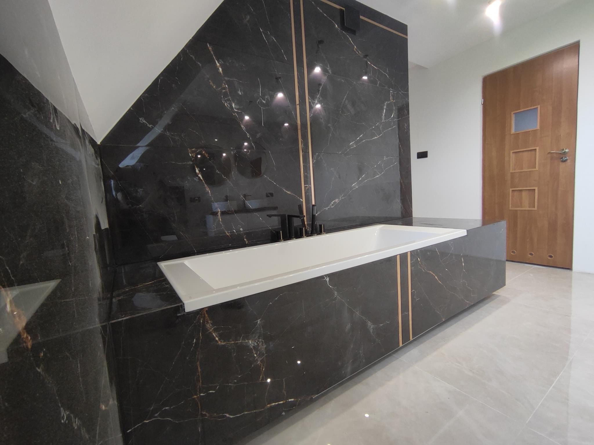 Nowoczesna łazienka z czarnymi marmurowymi płytkami z złotymi żyłami na ścianach i obudowie wanny, minimalistyczna armatura, jasne płytki podłogowe i drewniane drzwi z przeszkleniami w tle.