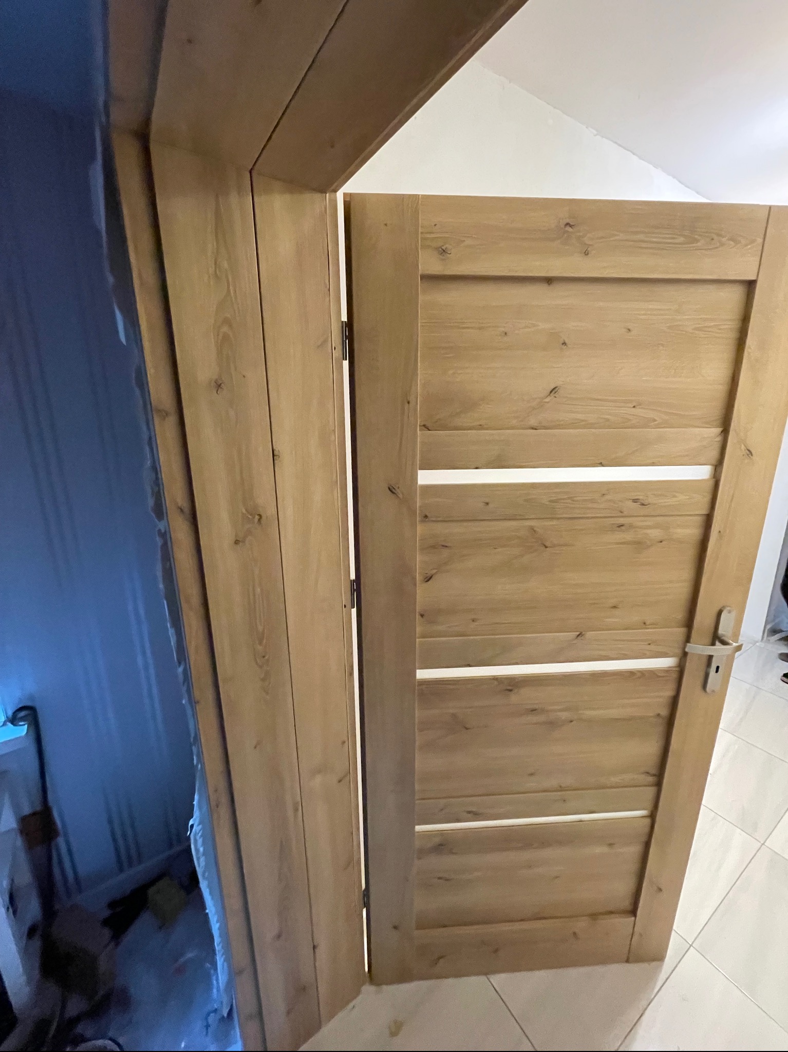 Otwarta, drewniana, wewnętrzna ościeżnica z zamontowanymi drzwiami z poziomymi, białymi intarsjami, widoczne elementy wykończeniowe ościeżnicy oraz fragment kafelek na podłodze.