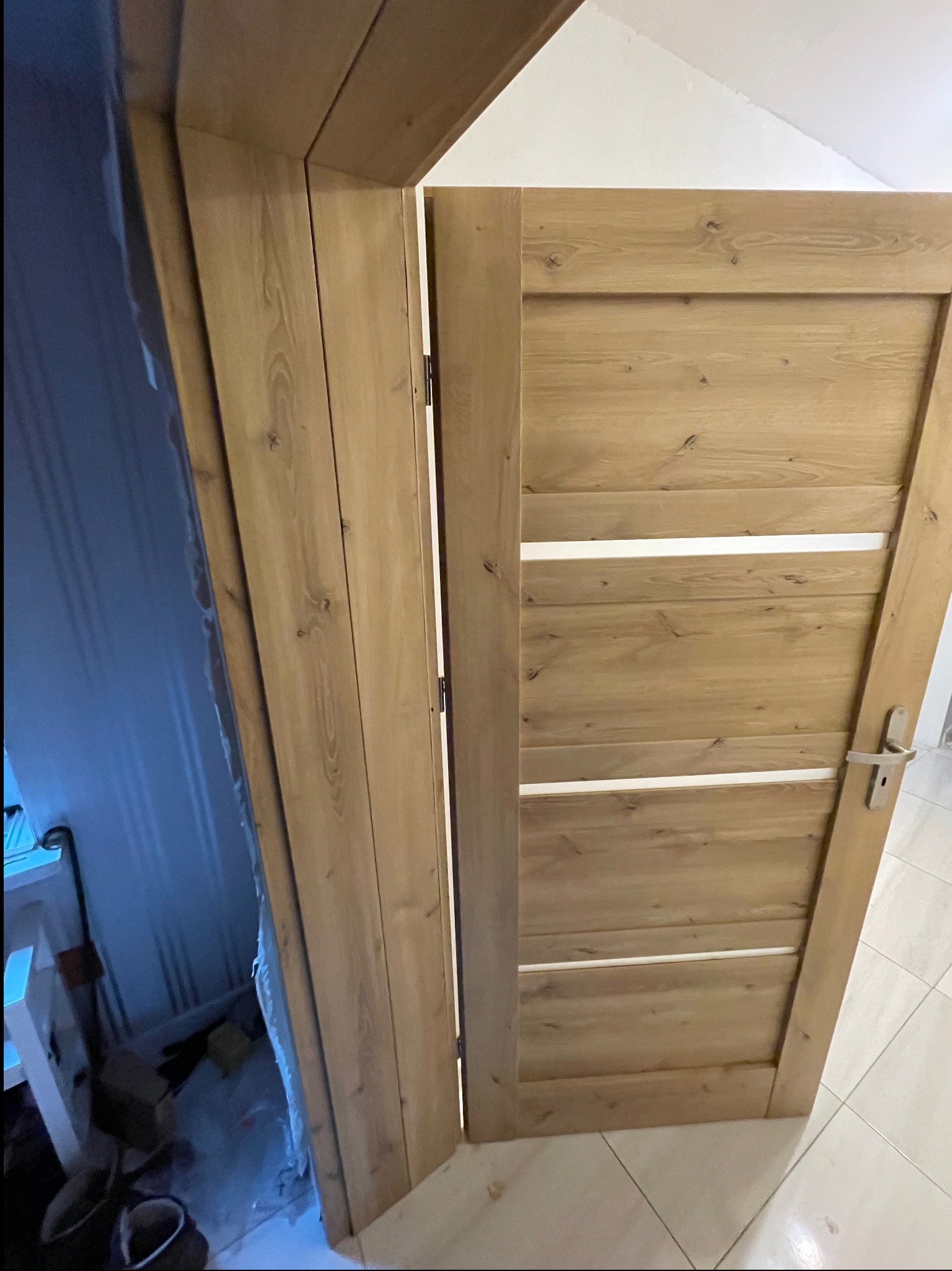 Otwarta, drewniana, wewnętrzna drzwi z trzema poziomymi, białymi wstawkami, widoczna ościeżnica i fragment pomieszczenia z niebieską ścianą oraz białymi meblami w tle.