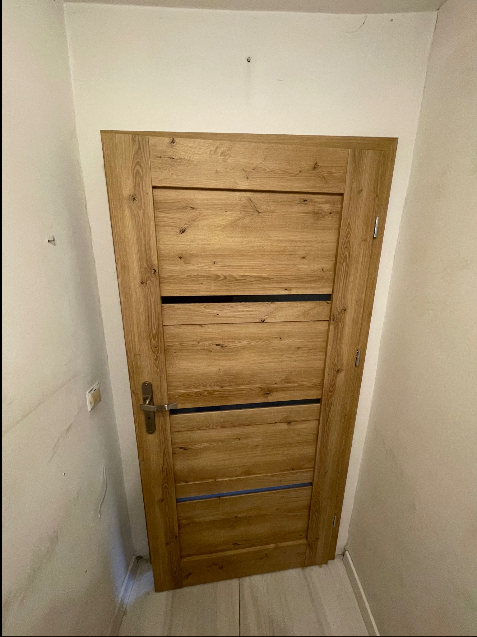 Zamknięte, drewniane drzwi wewnętrzne z poziomymi, czarnymi intarsjami, zamontowane w ościeżnicy w pomalowanym na biało pomieszczeniu z widocznymi niedoskonałościami na ścianach.