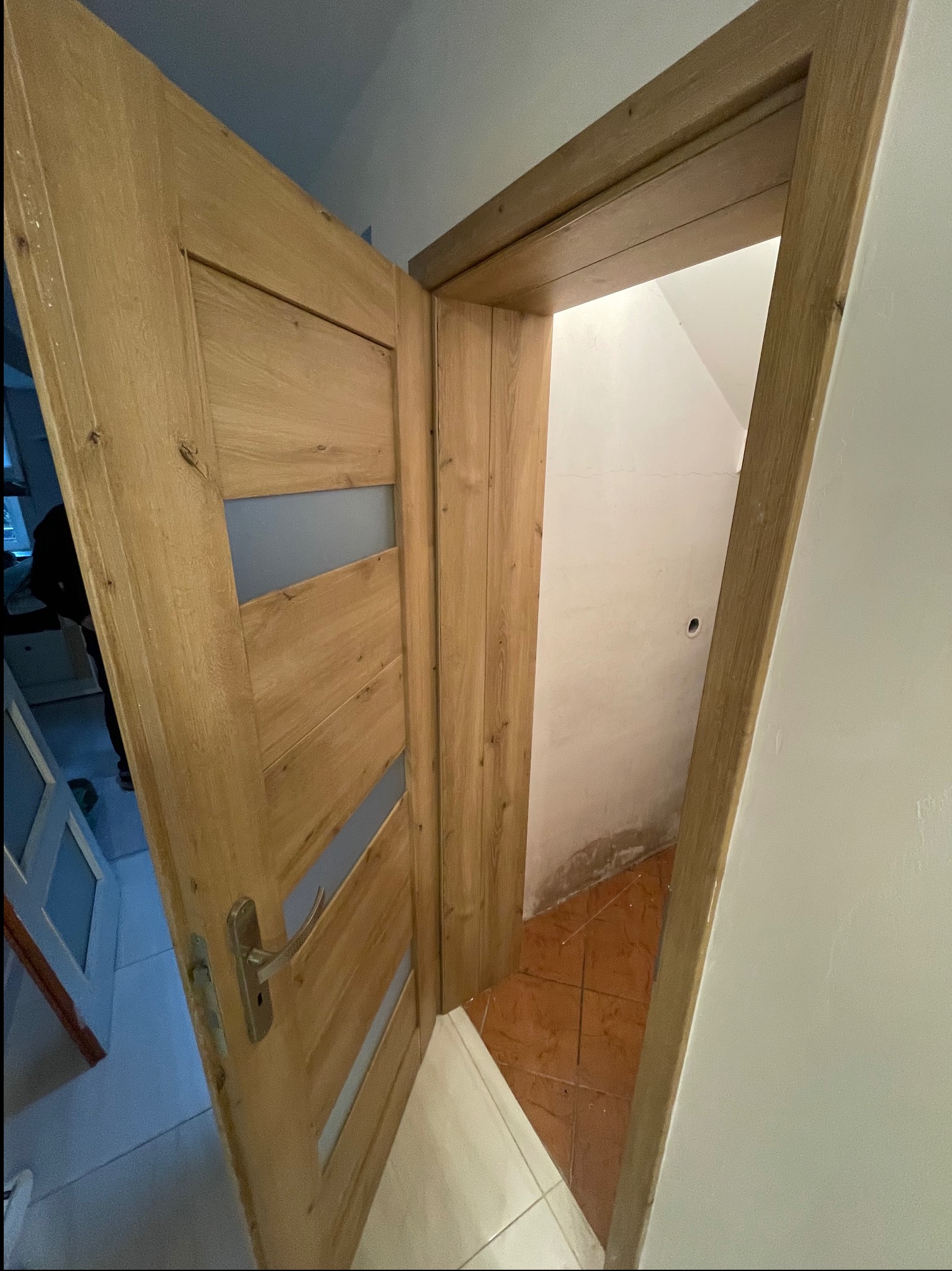 Otwarta drewniana dębowa drzwi z przeszkleniami, widoczny fragment korytarza z brązowymi płytkami na podłodze, białe ściany, widoczne ślady po remoncie przy podłodze.