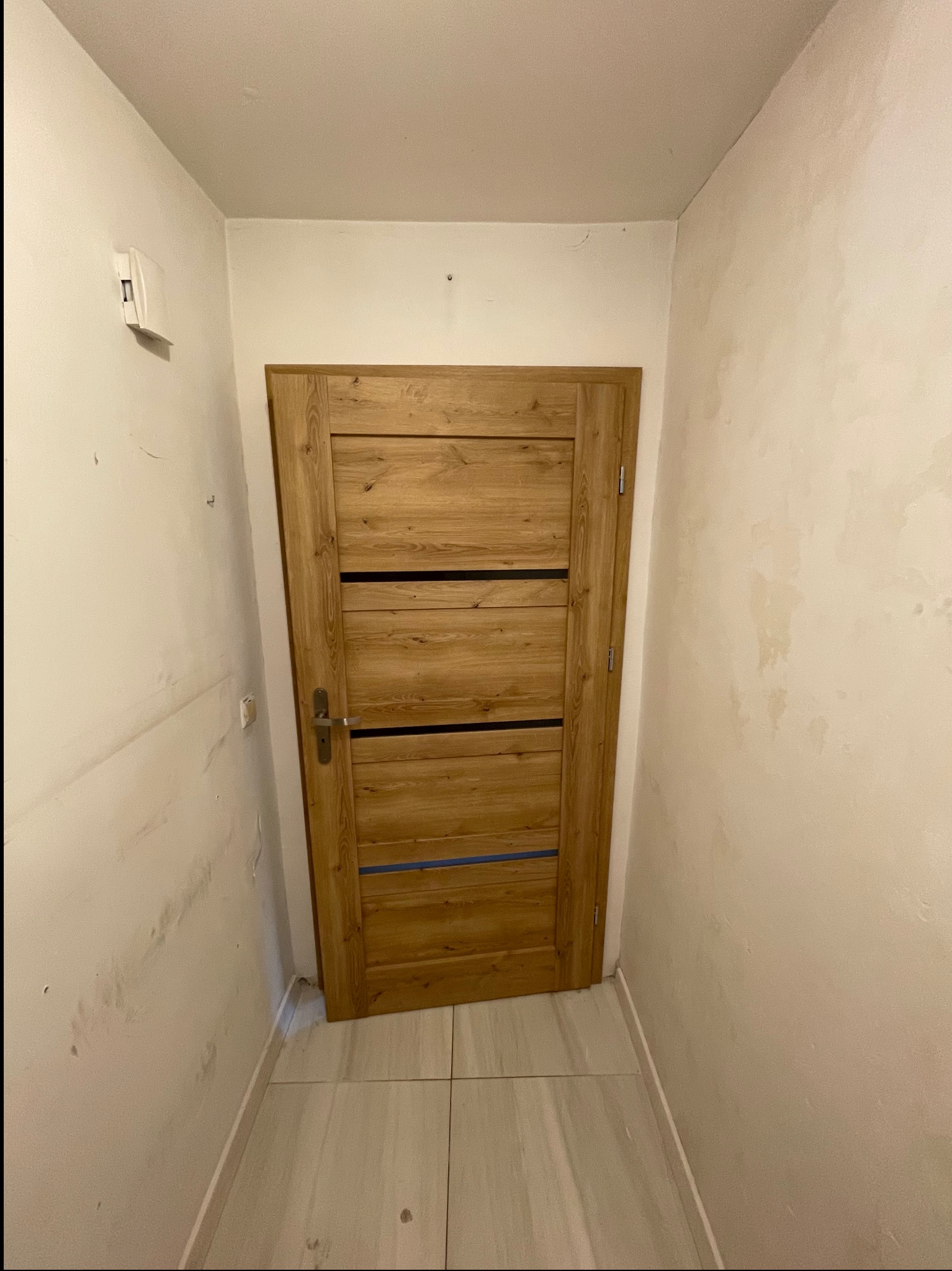 Zamknięte drewniane drzwi z trzema poziomymi czarnymi paskami w wąskim korytarzu z beżowymi ścianami i podłogą z płytek. Ściany wymagają odświeżenia.