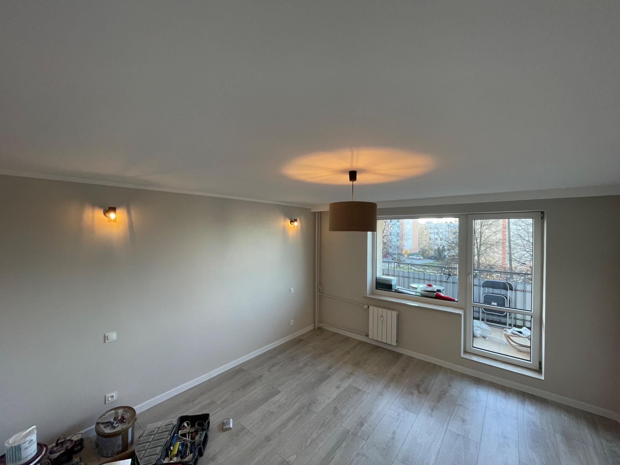 Pomalowane na jasny kolor wnętrze pokoju z panelami na podłodze, widoczne narzędzia malarskie i balkon z widokiem na zewnątrz.