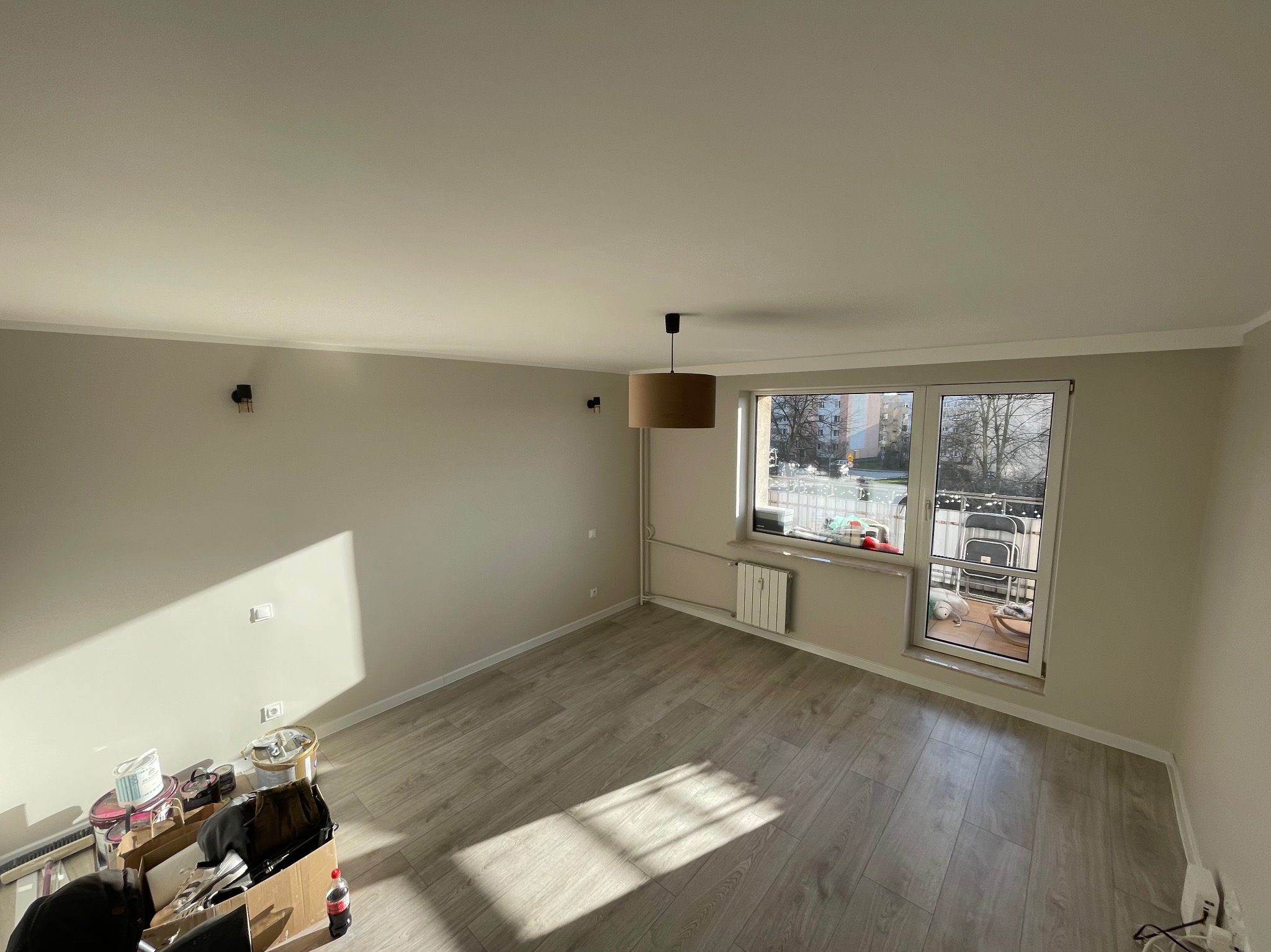 Pomalowane na beżowo puste pomieszczenie z panelami podłogowymi, widoczne puszki farb i narzędzia w rogu, okno balkonowe z widokiem na balkon.