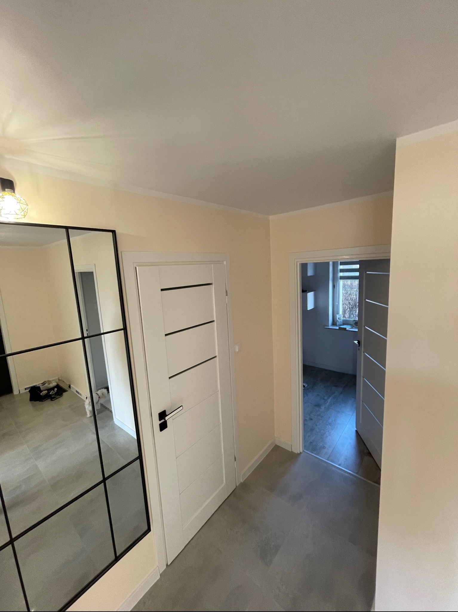 Wnętrze mieszkania po remoncie: białe drzwi z czarnymi poziomymi paskami i klamką, duże lustro z czarną ramą, szara podłoga, widok na kolejne pomieszczenie z otwartymi drzwiami.