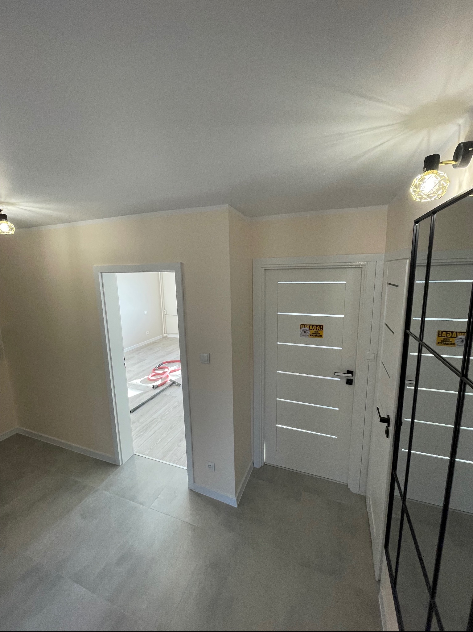 Nowoczesne wnętrze mieszkania po remoncie: szare płytki podłogowe, białe drzwi z poziomymi frezowaniami i naklejką ostrzegawczą, widoczne fragmenty zabudowy z czarnymi profilami, jasne ściany...