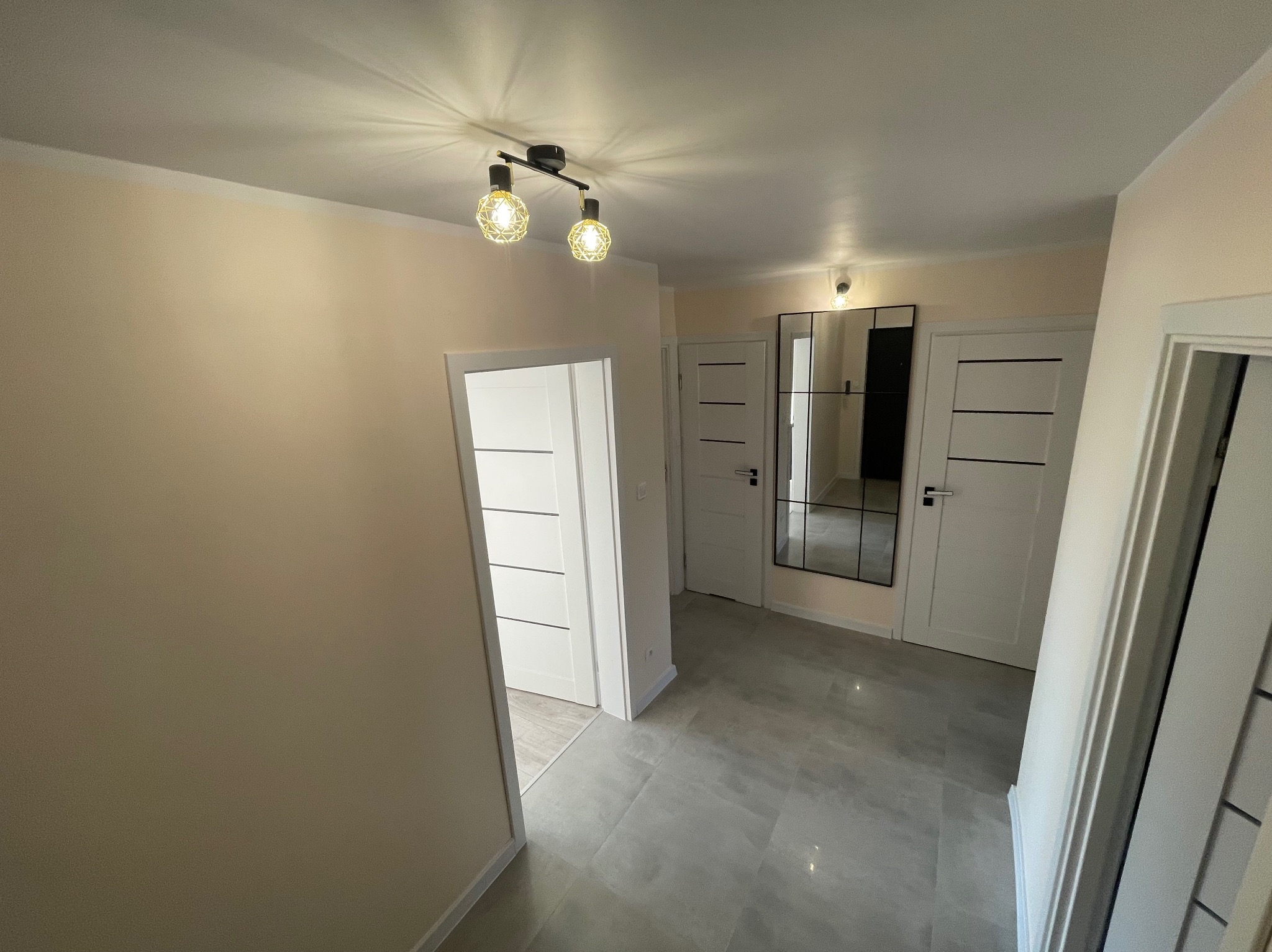 Jasne wnętrze korytarza z szarymi płytkami podłogowymi, białymi drzwiami z poziomymi frezami, lustrem w czarnej ramie i oświetleniem punktowym z ozdobnymi żarówkami.